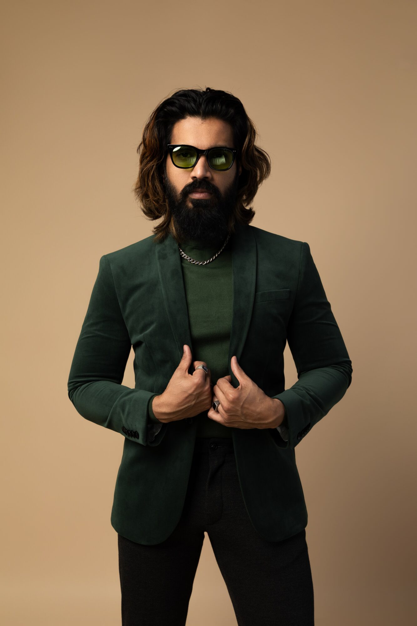Man with long hair, beard, sunglasses, wearing a dark green blazer and shirt, holding the blazer lapels, against a beige background.