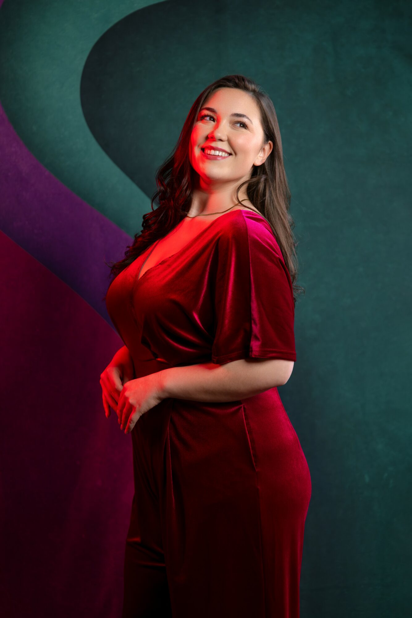 Smiling woman in red velvet dress stands against a colorful abstract background.