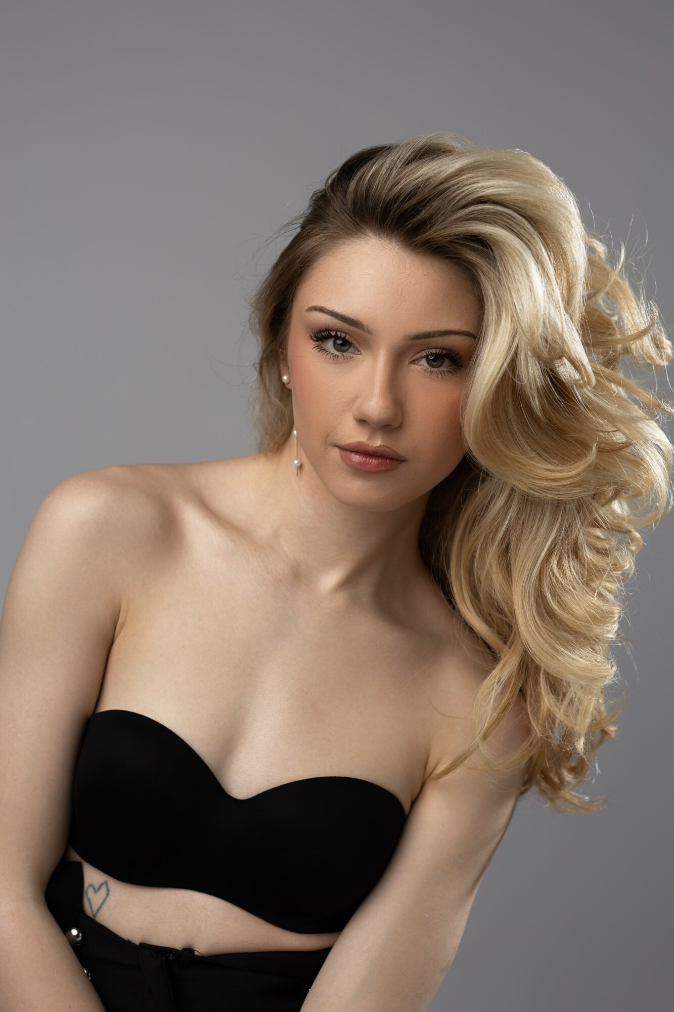 Young woman with long, wavy blonde hair and fair skin, wearing a black strapless top, against a gray background.