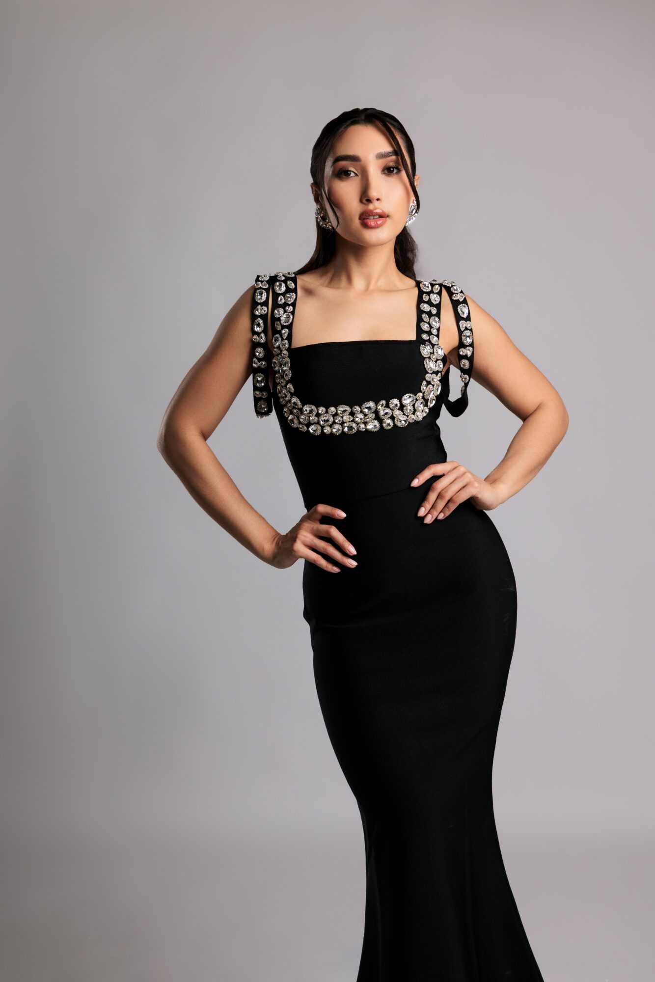 Woman in a black dress with embellished shoulder straps, standing with hands on hips, facing forward.