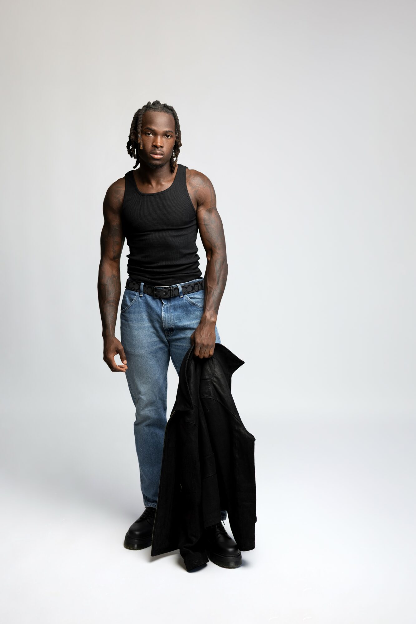Young man with dreadlocks wearing a black sleeveless shirt and jeans, holding a black jacket, standing against a plain background.