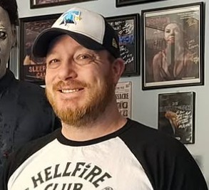 Man with a beard wearing a baseball cap and a black and white shirt, smiling, in front of framed pictures on a wall.