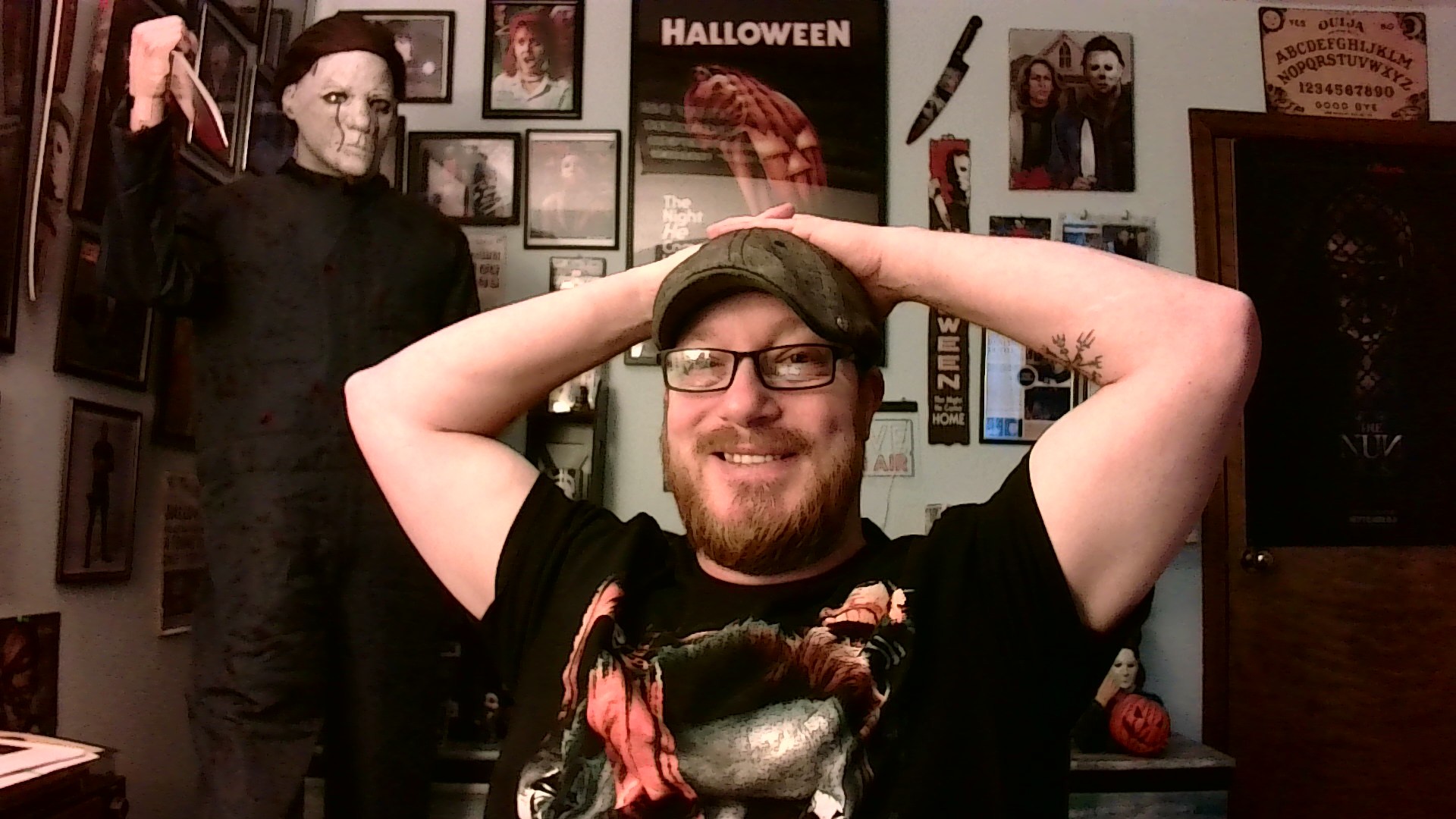Person smiling with glasses and a cap, arms raised, in a room decorated with Halloween posters and figures.