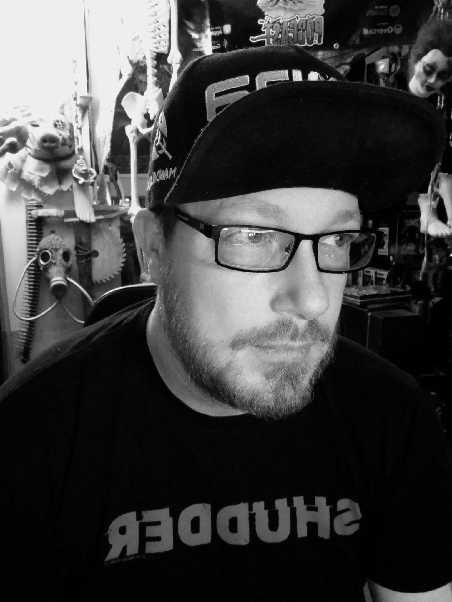 Man with glasses and a beard wearing a cap, sitting indoors with shelves and toys in the background.