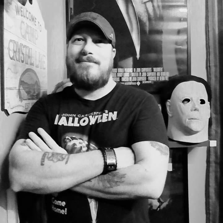 Man with beard and cap standing with arms crossed next to a mask of a face, in a room with posters and brick wall.