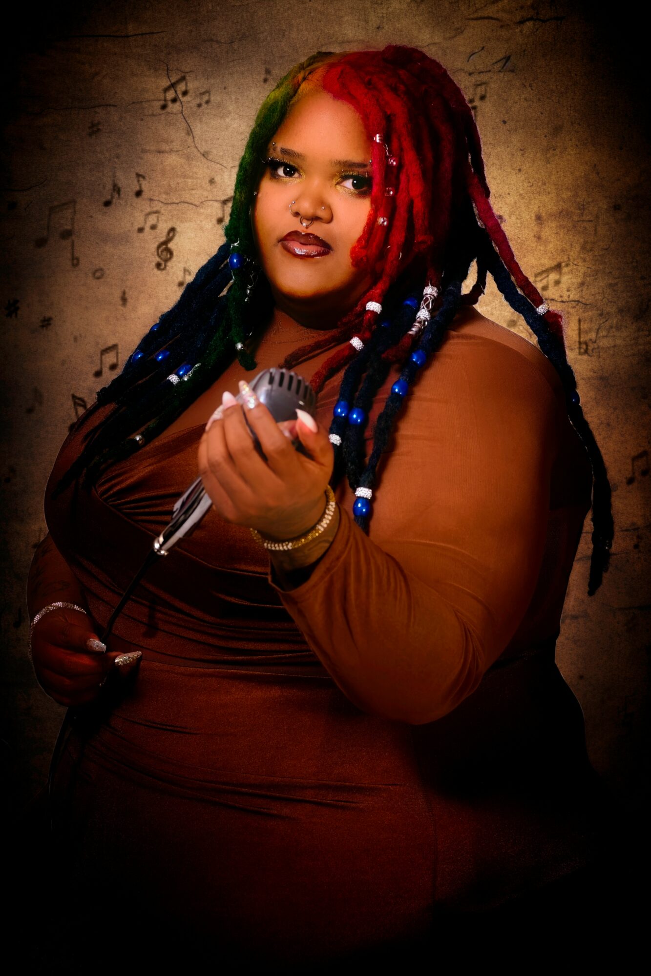Woman with colorful dreadlocks holding a microphone, wearing a brown top, against a textured background.