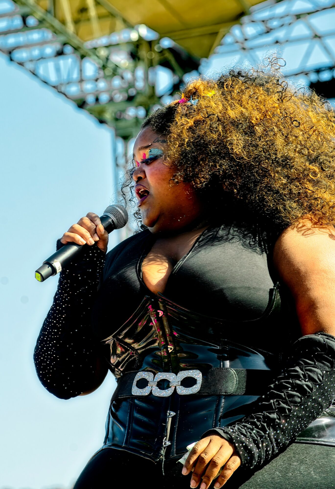A woman with curly hair singing into a microphone on stage, wearing a black outfit with glittery sleeves.