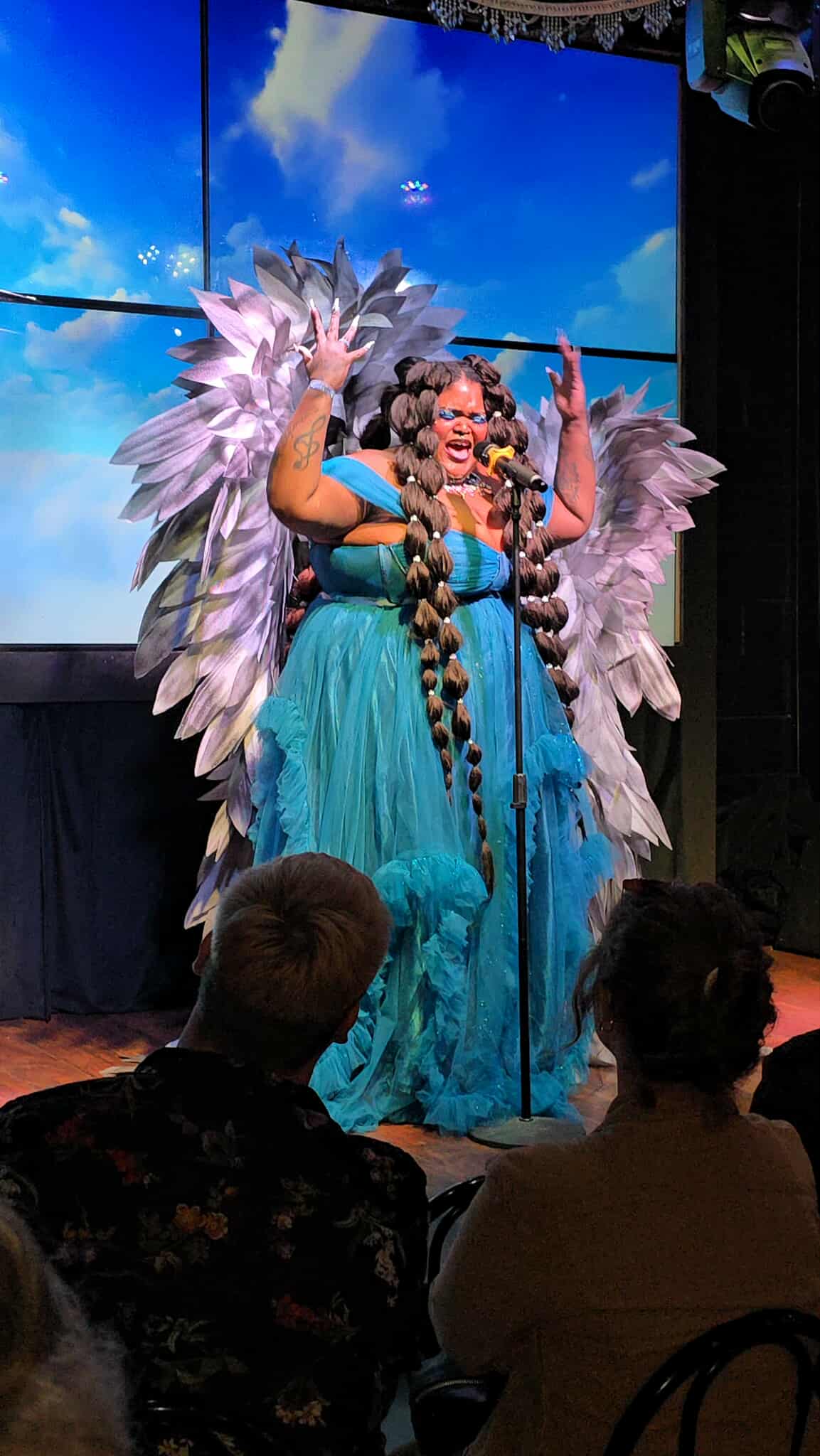 Performer in blue costume with large feathered wings on stage, smiling, with audience in foreground.