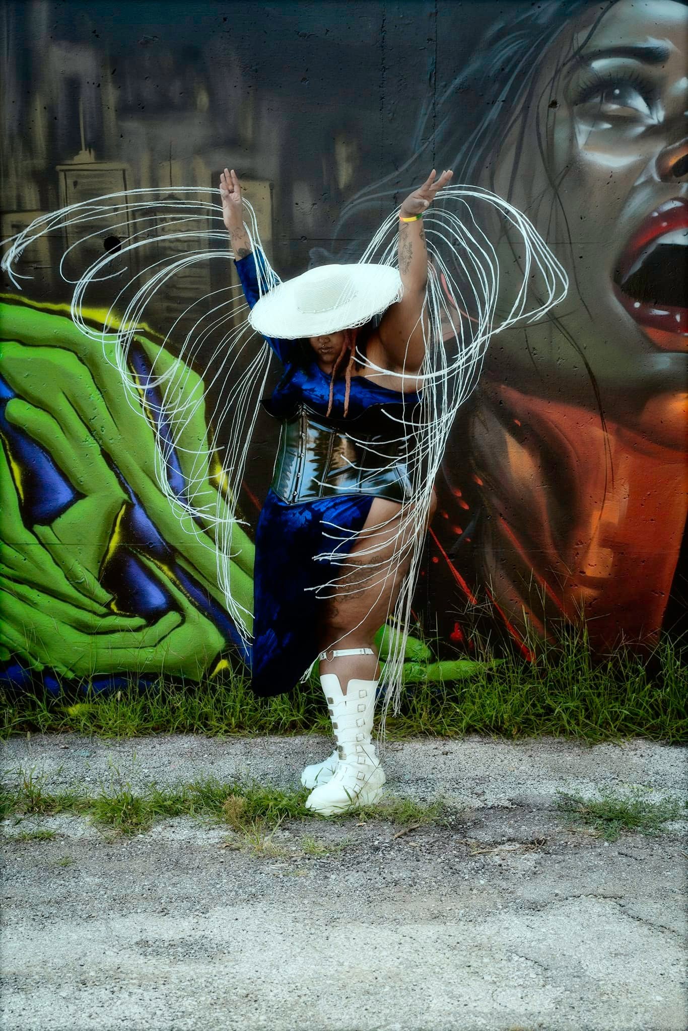 Person in costume with white hat and white boots standing in front of colorful graffiti mural, arms raised, on a grassy area.