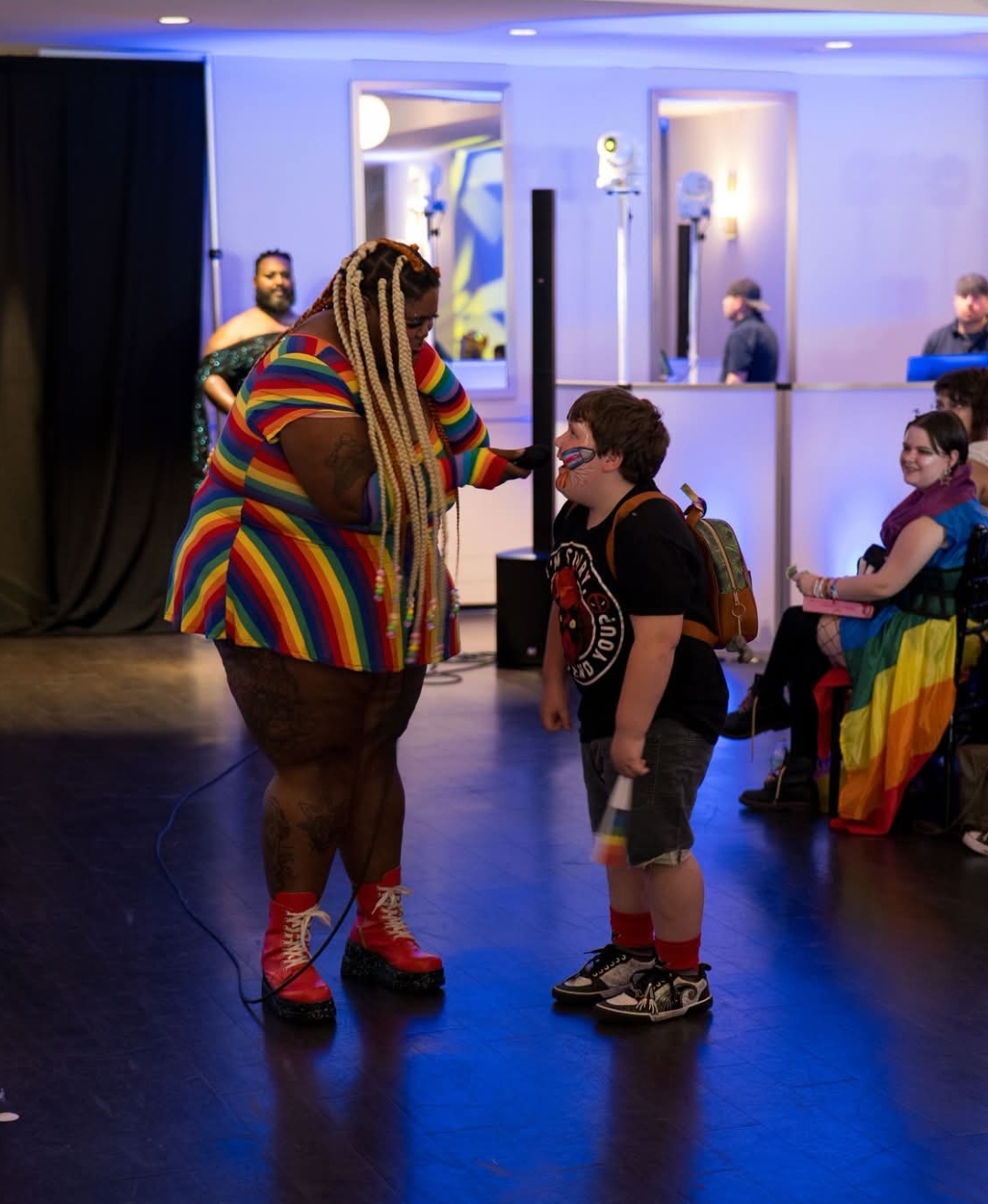 A performer with colorful striped clothing and long hair interacts with a young boy in a room with blue lighting.