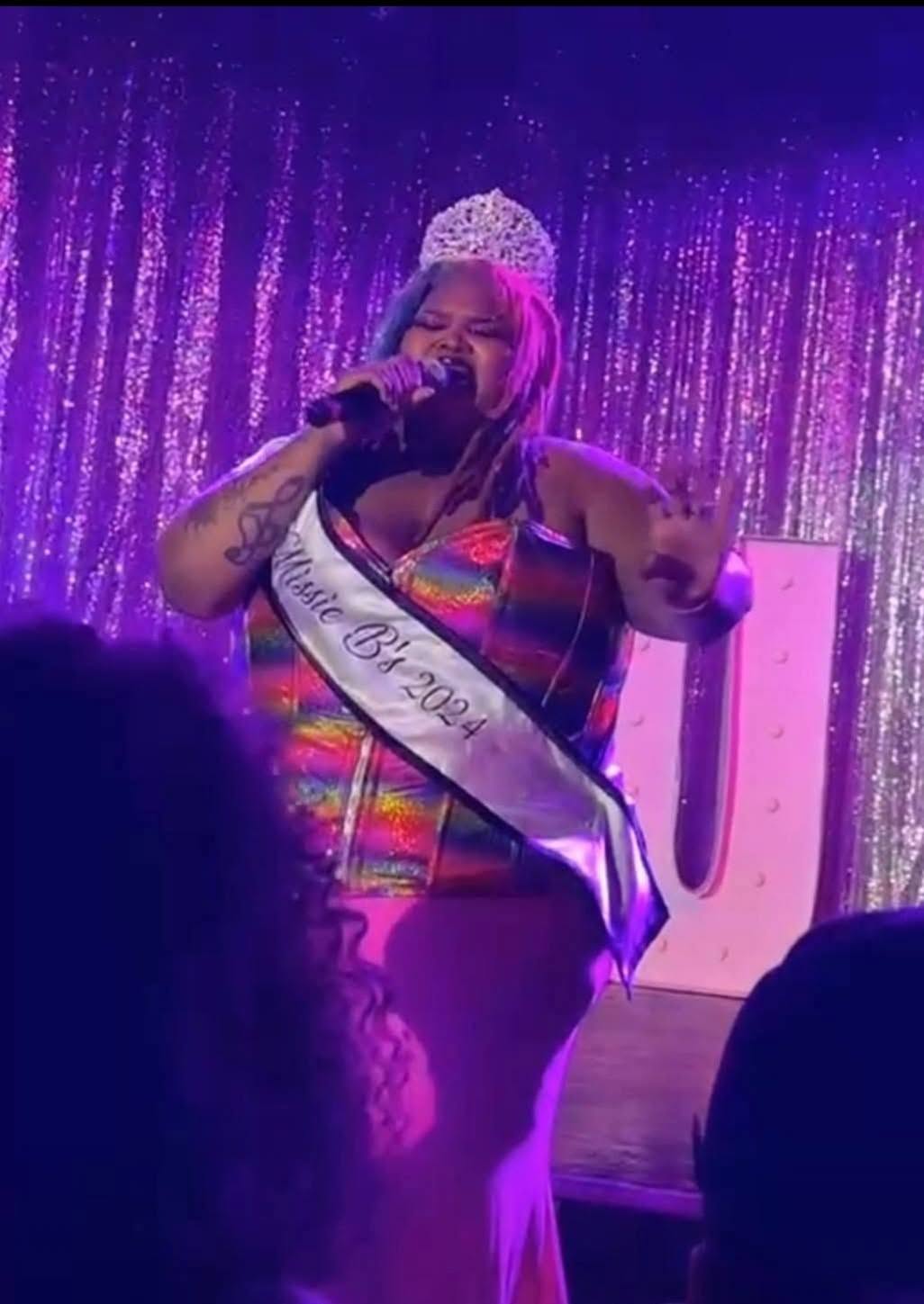 Performer with pink hair, wearing a tiara and sash, singing into a microphone on stage with purple curtains.