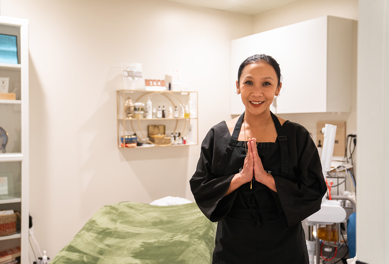 Woman in black uniform smiling with hands pressed together in a room with a bed and shelves.