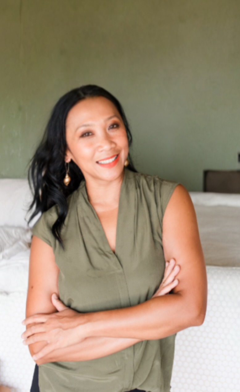 Woman with black hair smiling, arms crossed, wearing a green sleeveless top, standing indoors against a green wall.