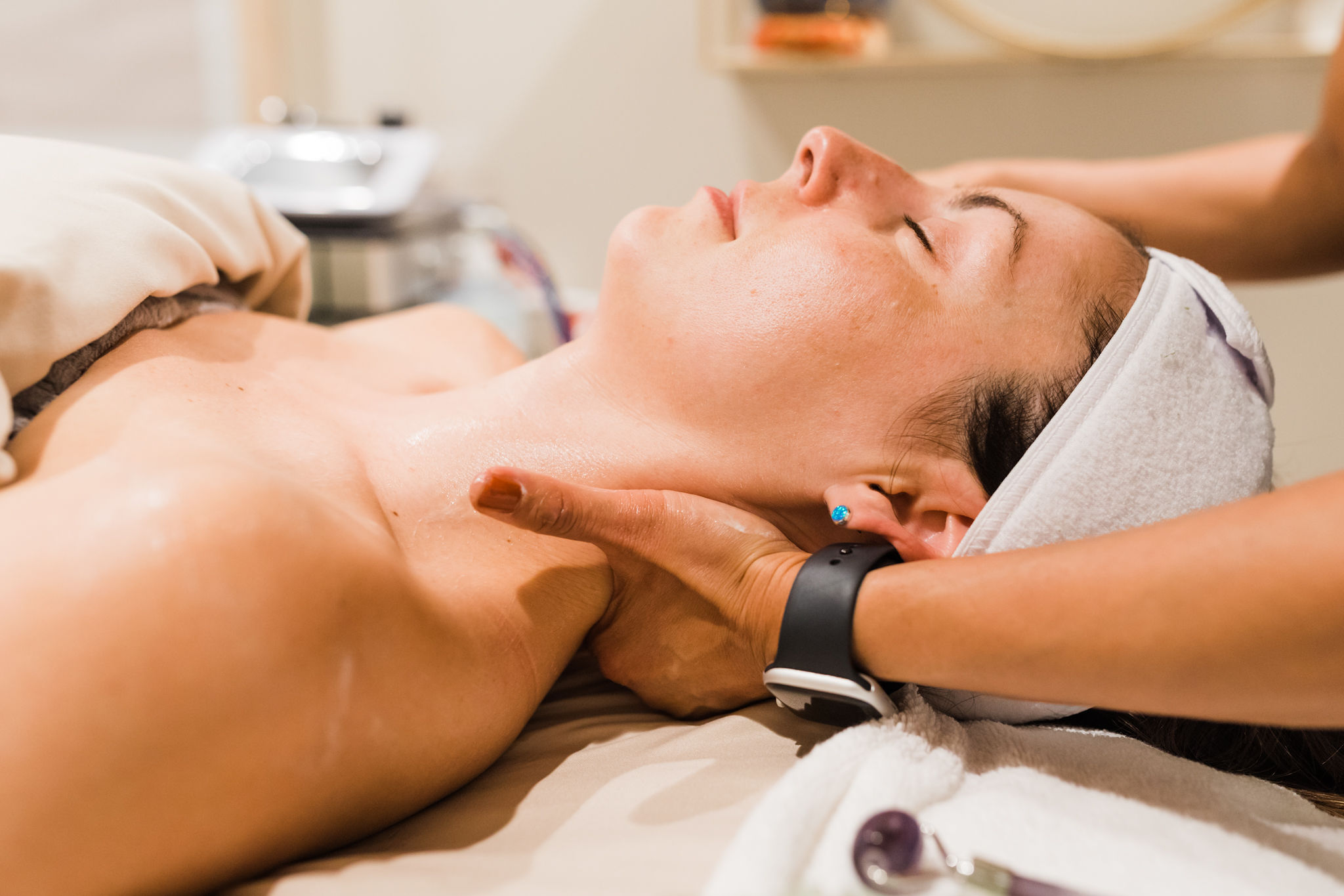 Person receiving facial treatment with eyes closed, lying on a bed, head supported by a therapist's hand, in a spa or clinic setting.