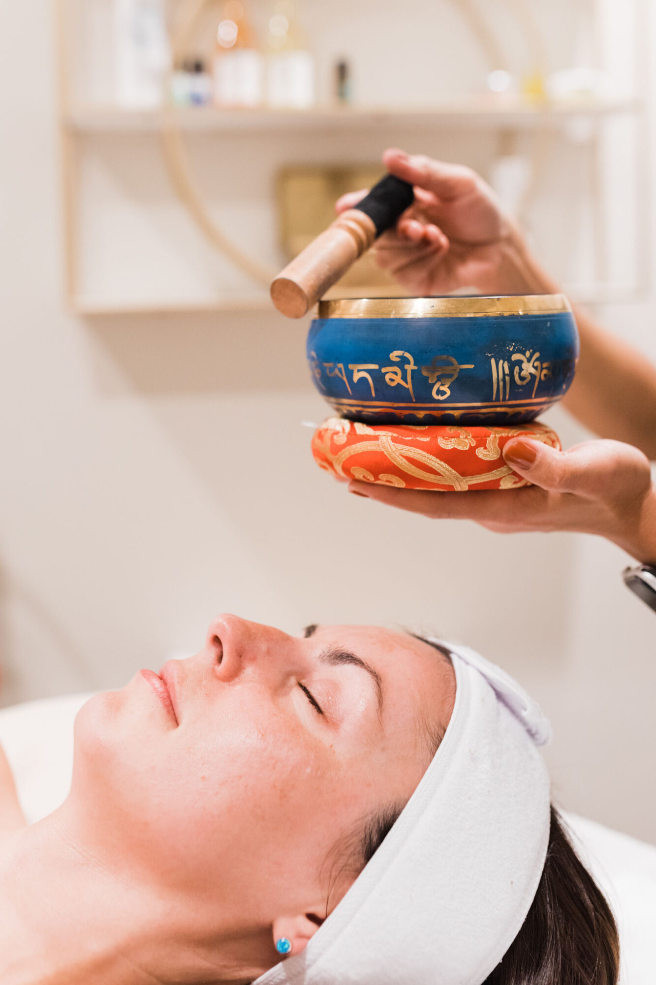 Person lying down with eyes closed, wearing a white headband, receiving a sound therapy session with bowls and a mallet.
