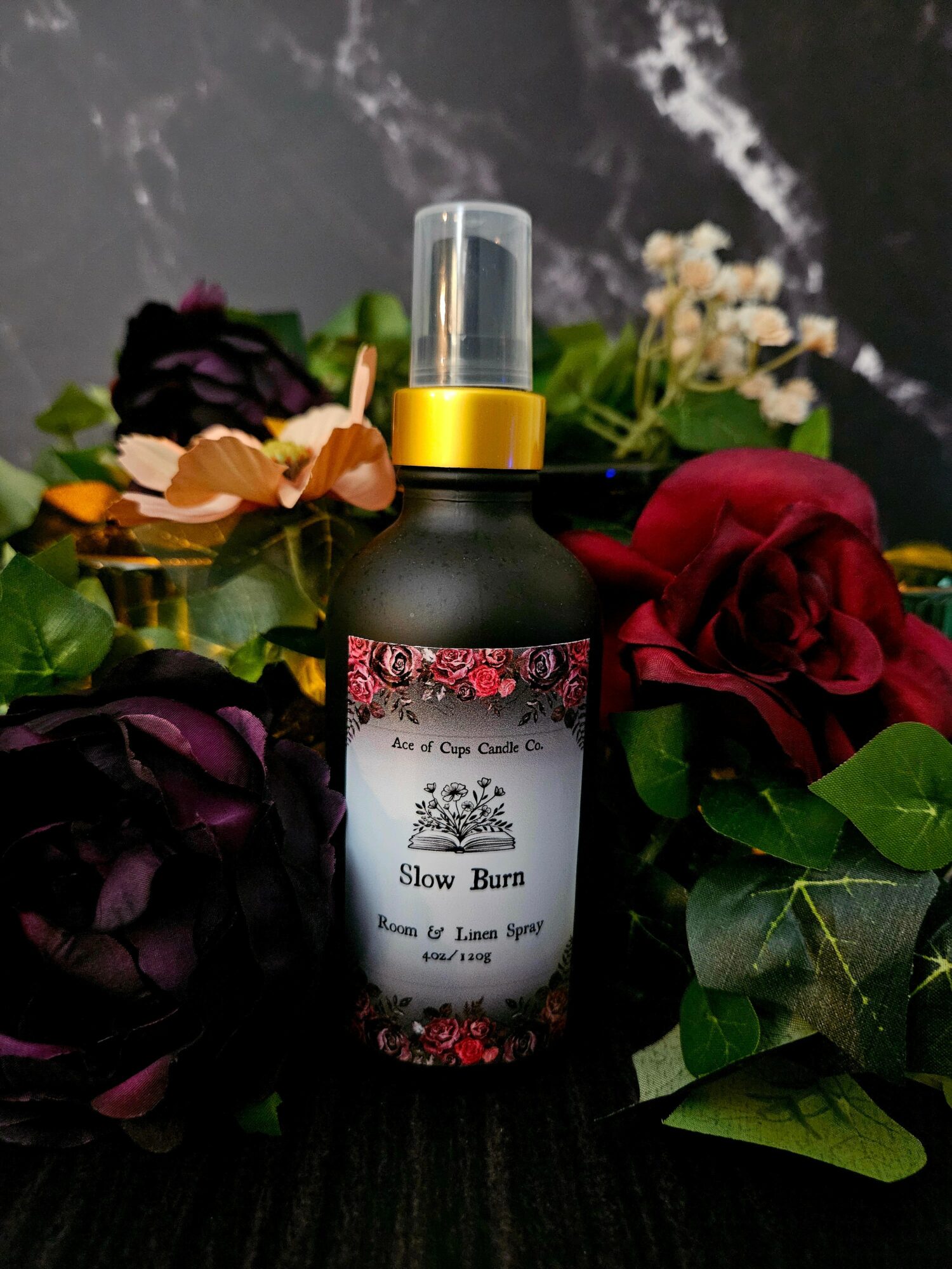 Black spray bottle with a floral label, surrounded by dark purple and red roses and green leaves, on a dark surface.