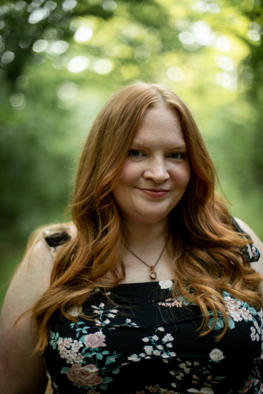 Young woman with long, wavy red hair smiling outdoors with green trees in background.