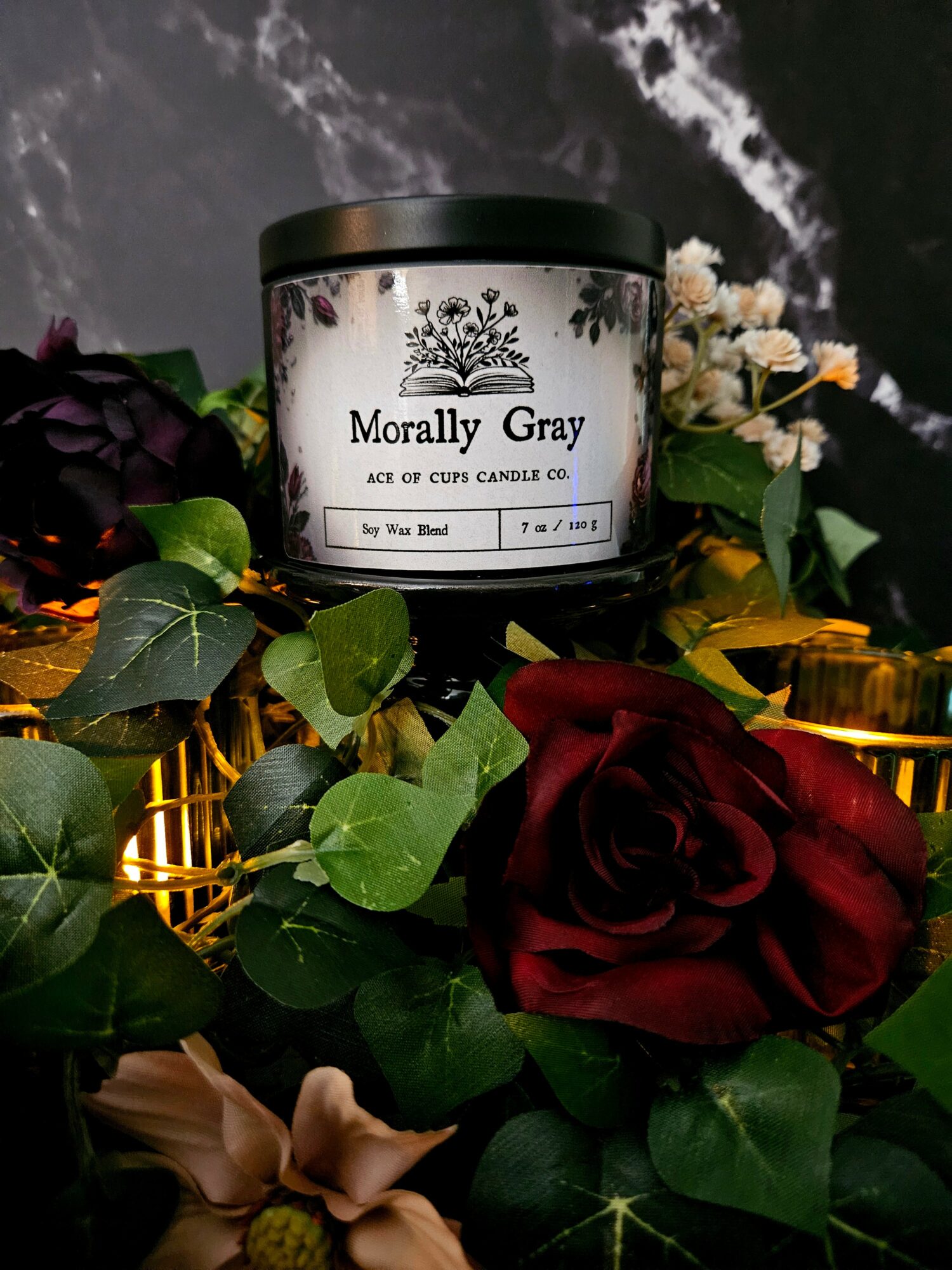 A black jar labeled 'Morally Gray' surrounded by red and pink roses and green leaves, with a dark marble background.
