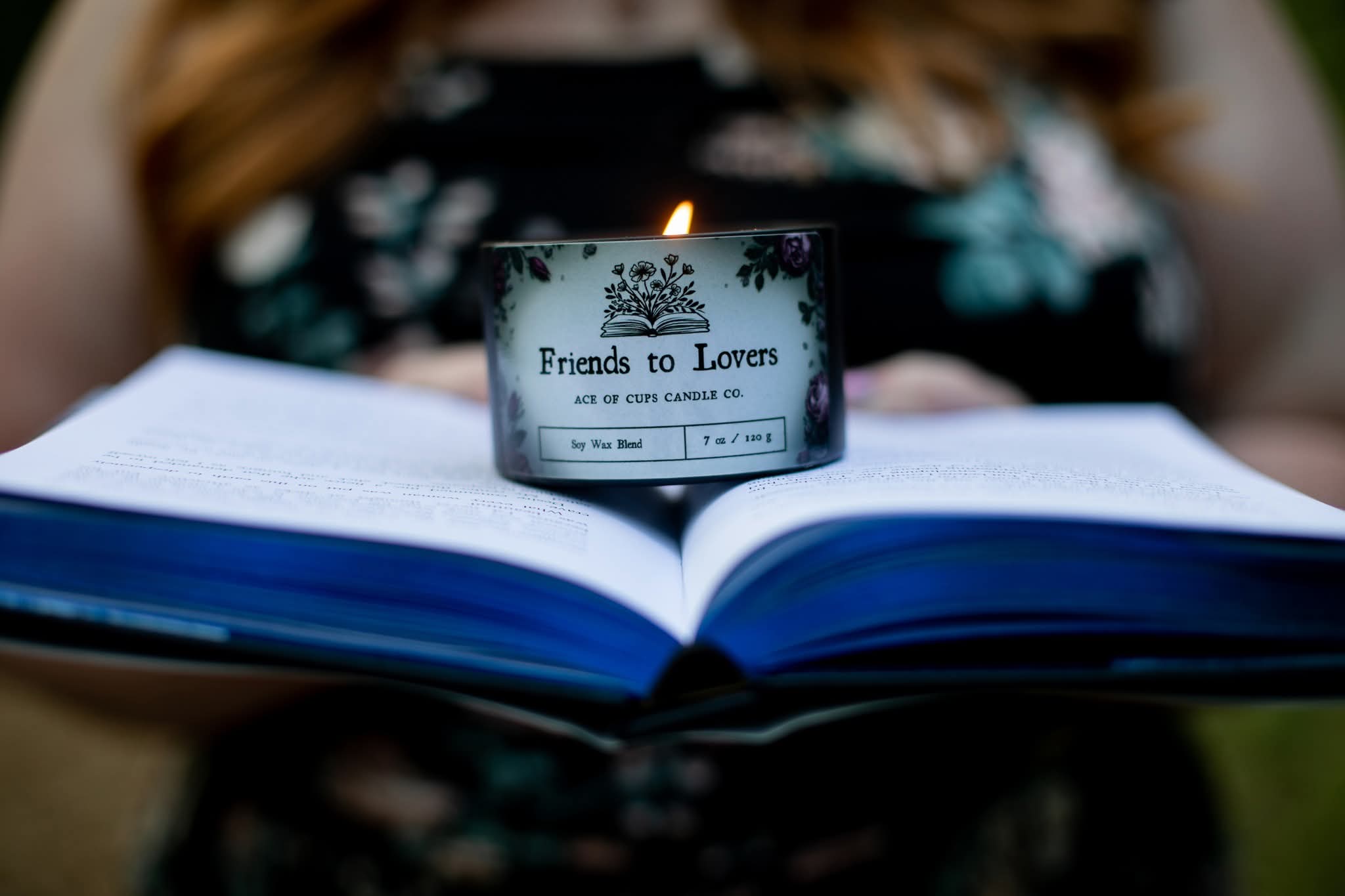 Open book with a small candle on top, labeled 'Friends to Lovers,' held by a person wearing a floral dress.