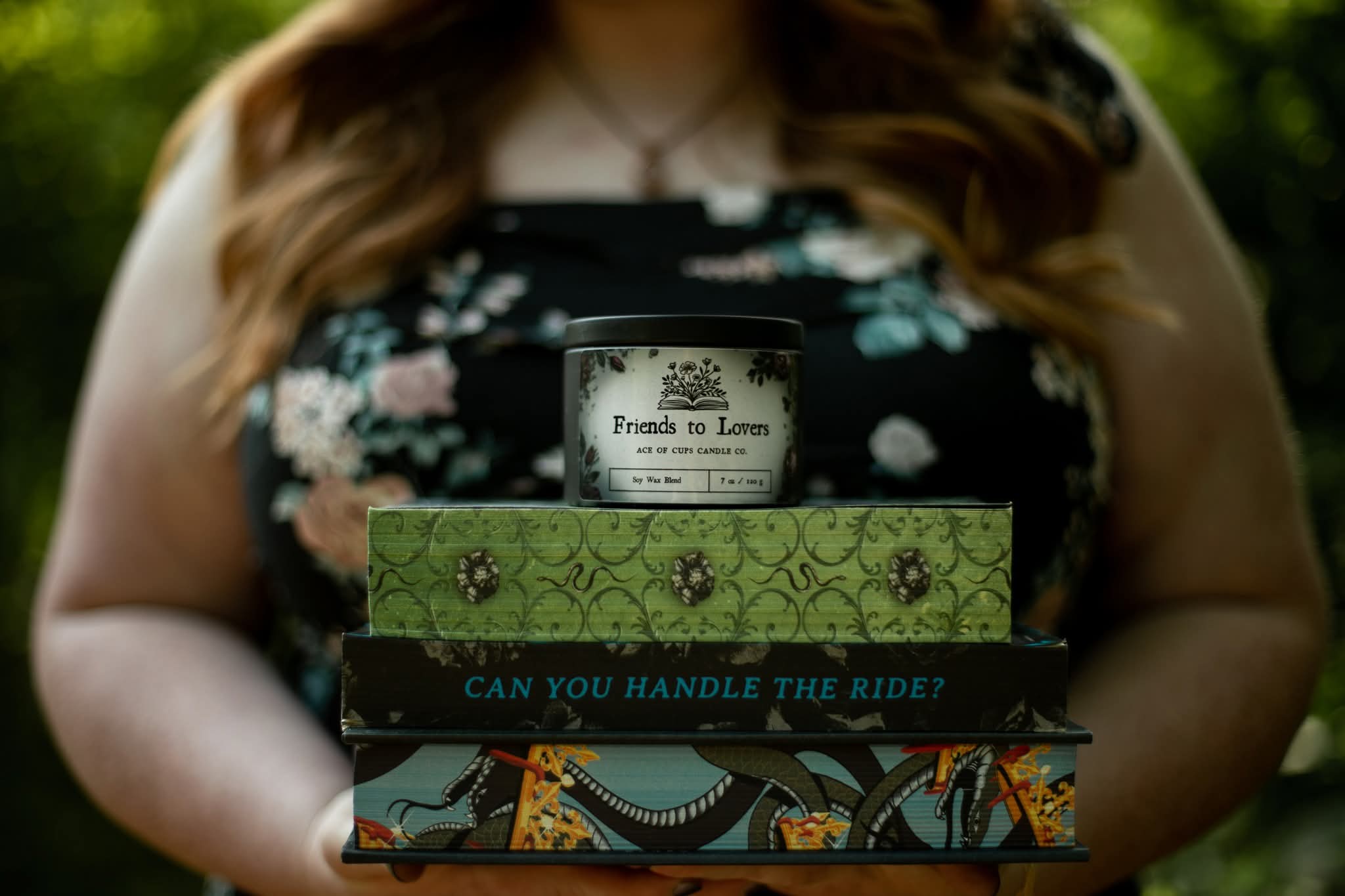 Person holding three books, with a blurred background of green foliage, wearing a floral top, face not visible.