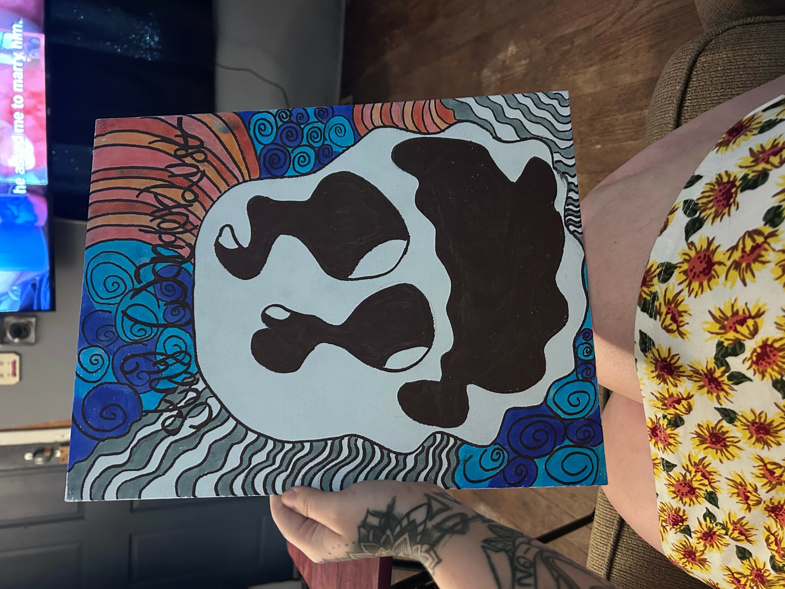 Colorful abstract painting with black shapes on a light background, surrounded by blue and orange patterns, held by a person with a tattooed hand.