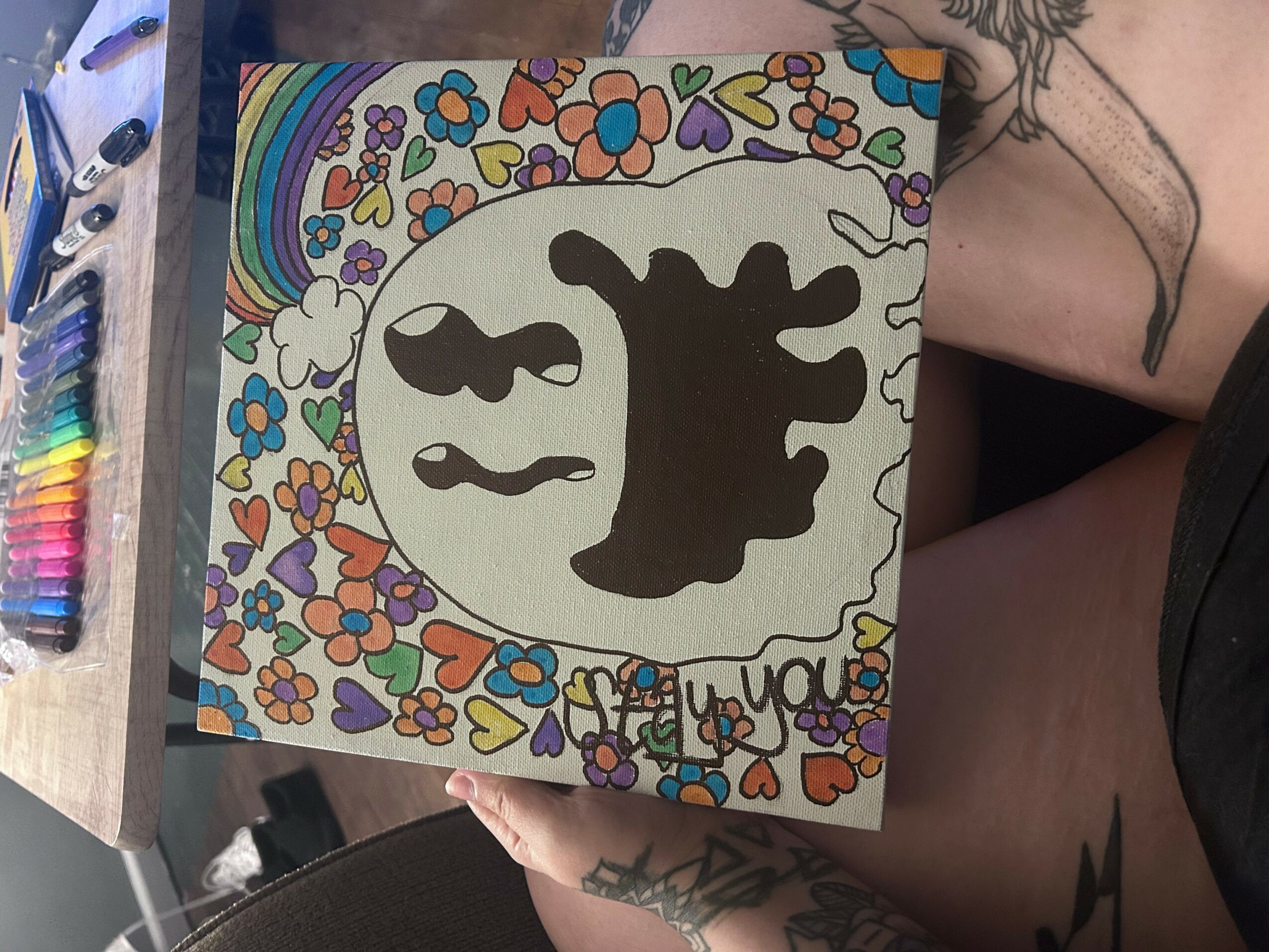 Colorful drawing with flowers, rainbow, and a black silhouette of a creature, with handwritten text at the bottom.
