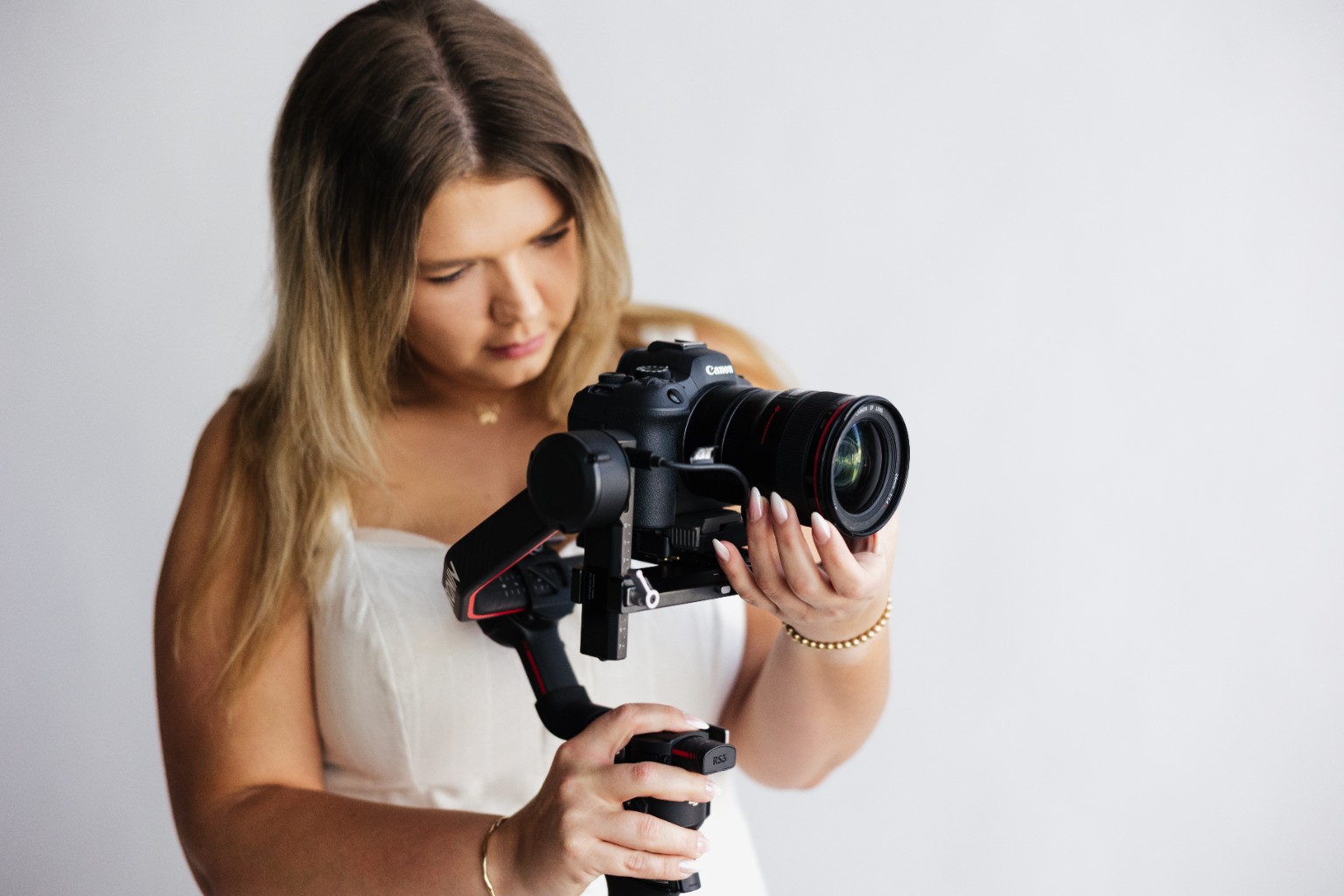 Young woman with long hair holding a camera, looking down at it, against a plain background.
