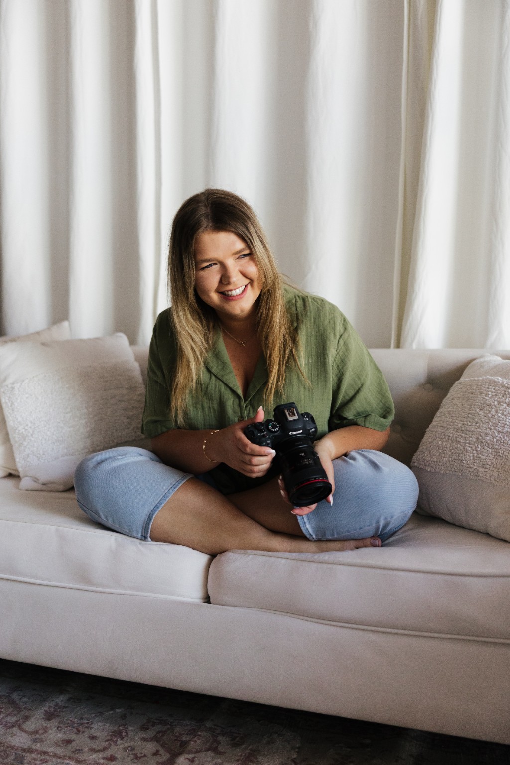 Young woman sitting on a sofa, smiling, holding a camera, with white curtains behind her.
