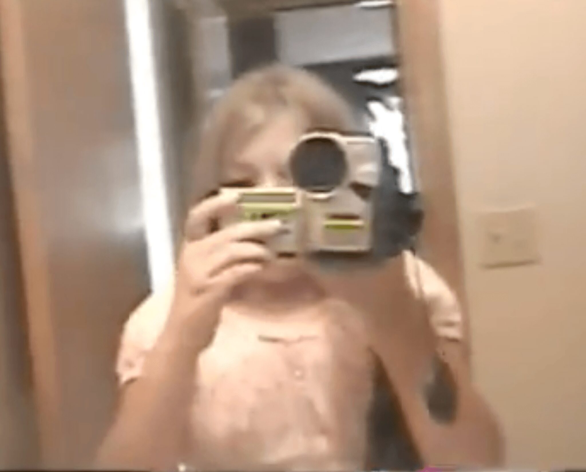 Person taking a mirror selfie with a camera, wearing a light-colored top, in an indoor setting.