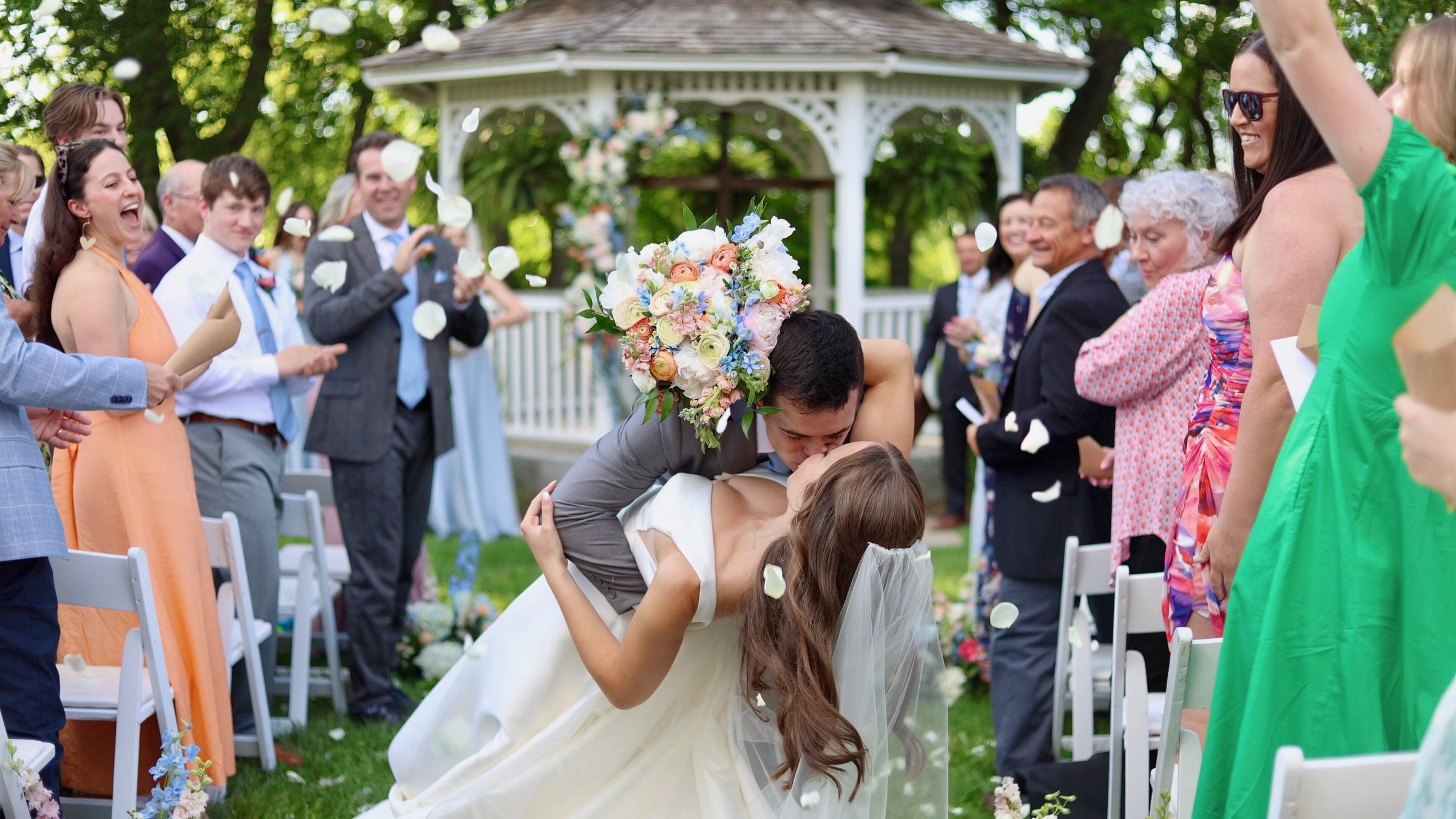 Bride and groom kiss outdoors surrounded by guests, with a floral bouquet and gazebo in the background.
