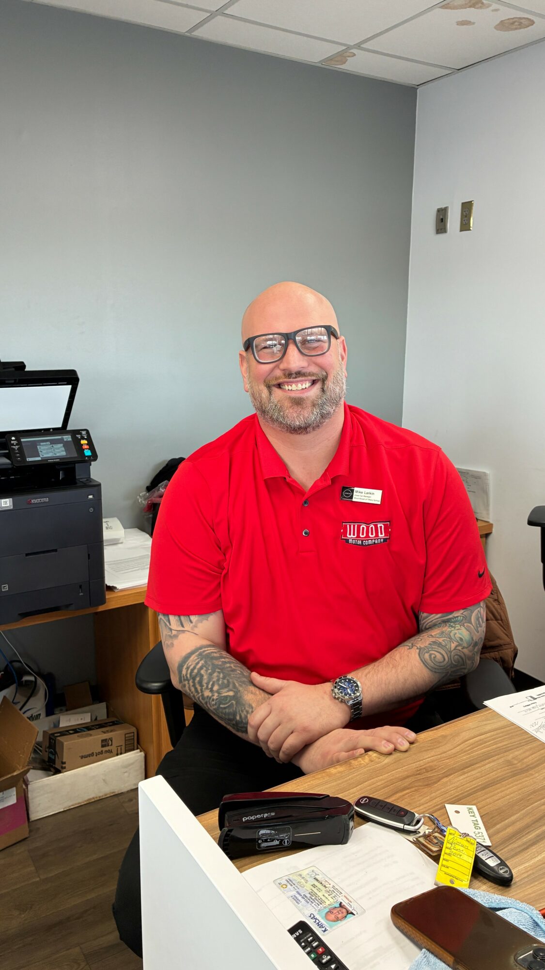 Man with glasses and tattoos wearing a red shirt, smiling, sitting at a desk in an office.
