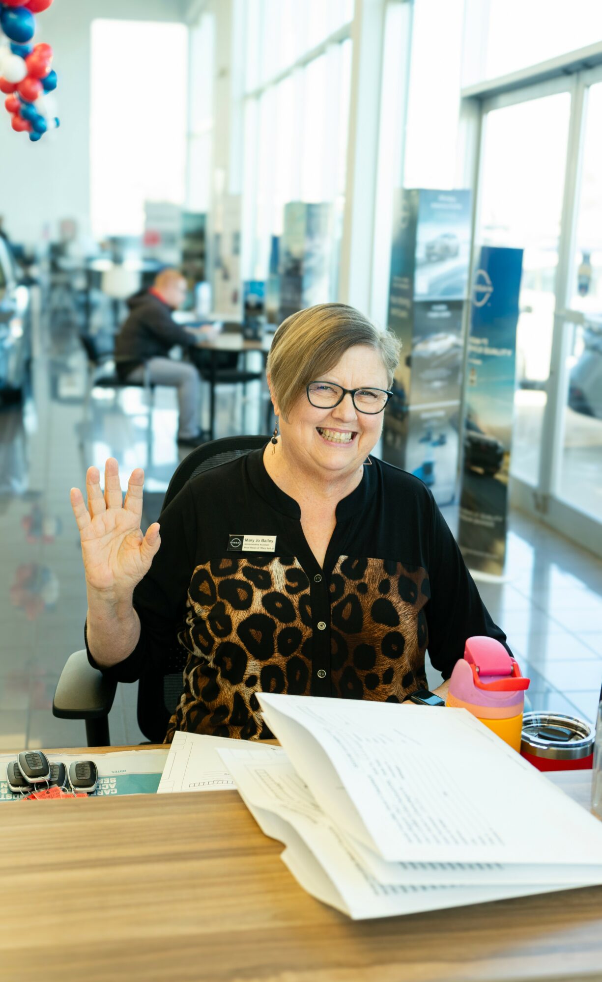 Smiling woman with glasses waving at camera, seated at a desk in a bright airport terminal, with papers and a pink water bottle.