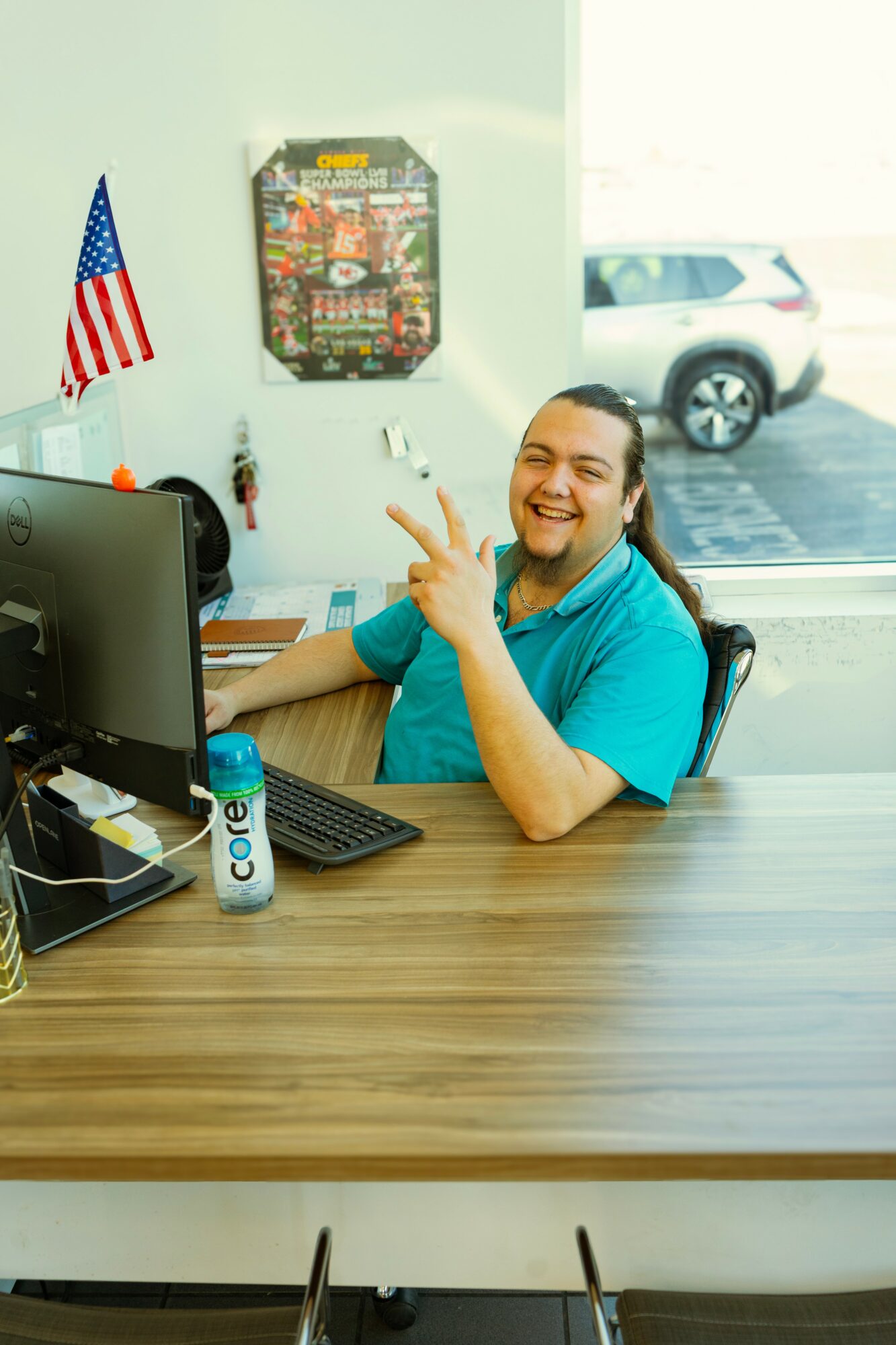 Person sitting at desk smiling, pointing, with computer, soda can, and American flag in background.