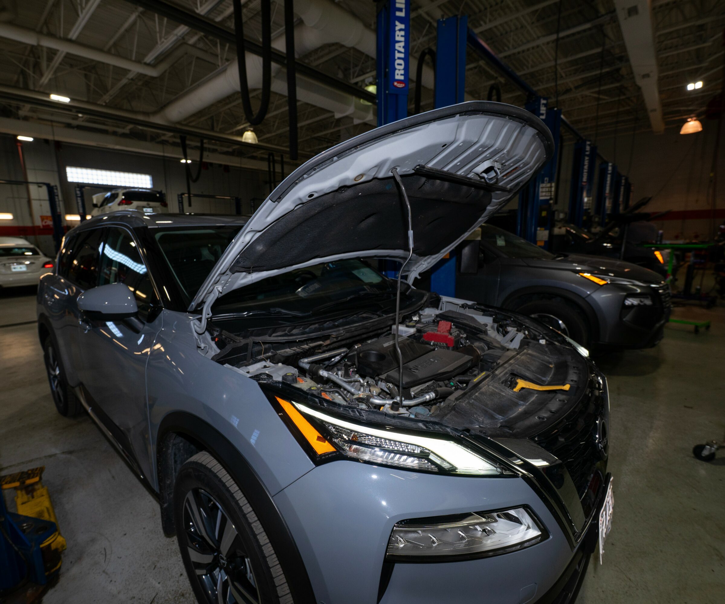 Gray car with open hood inside a garage, engine visible, surrounded by other vehicles and equipment.