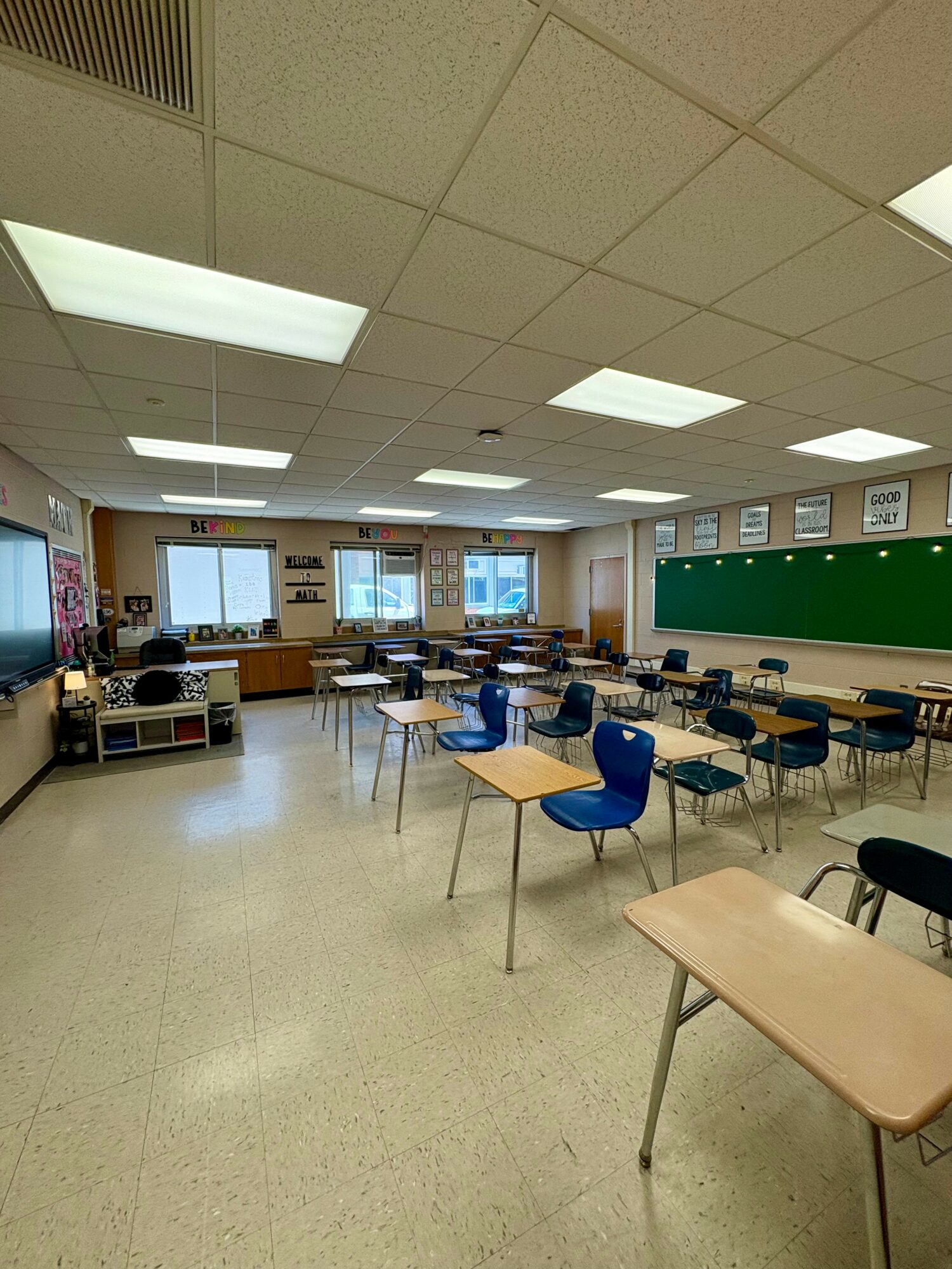 Empty classroom with desks and chairs, green chalkboard, windows, and ceiling lights.