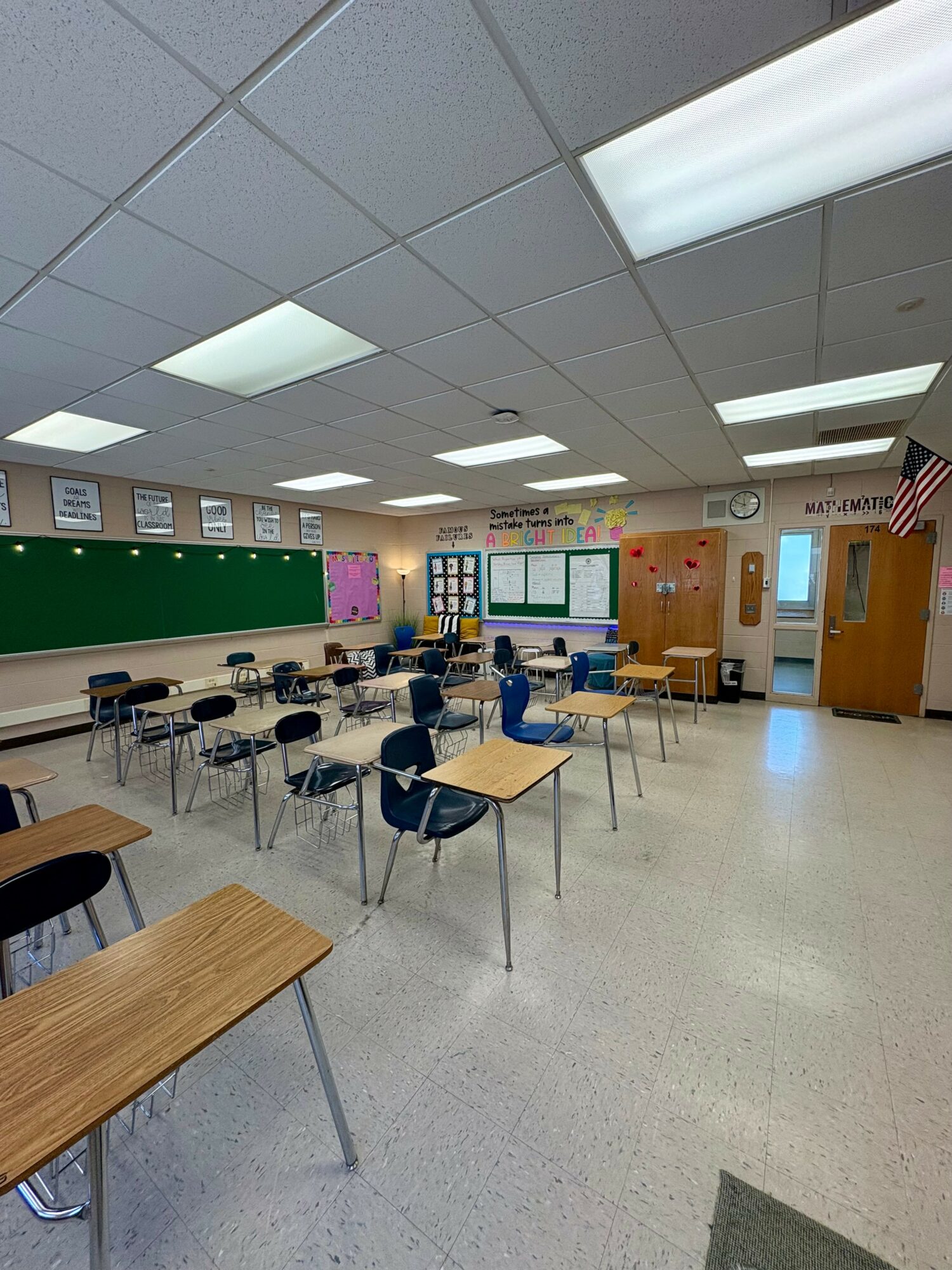Empty classroom with desks, chairs, a green chalkboard, and bulletin boards, illuminated by ceiling lights.