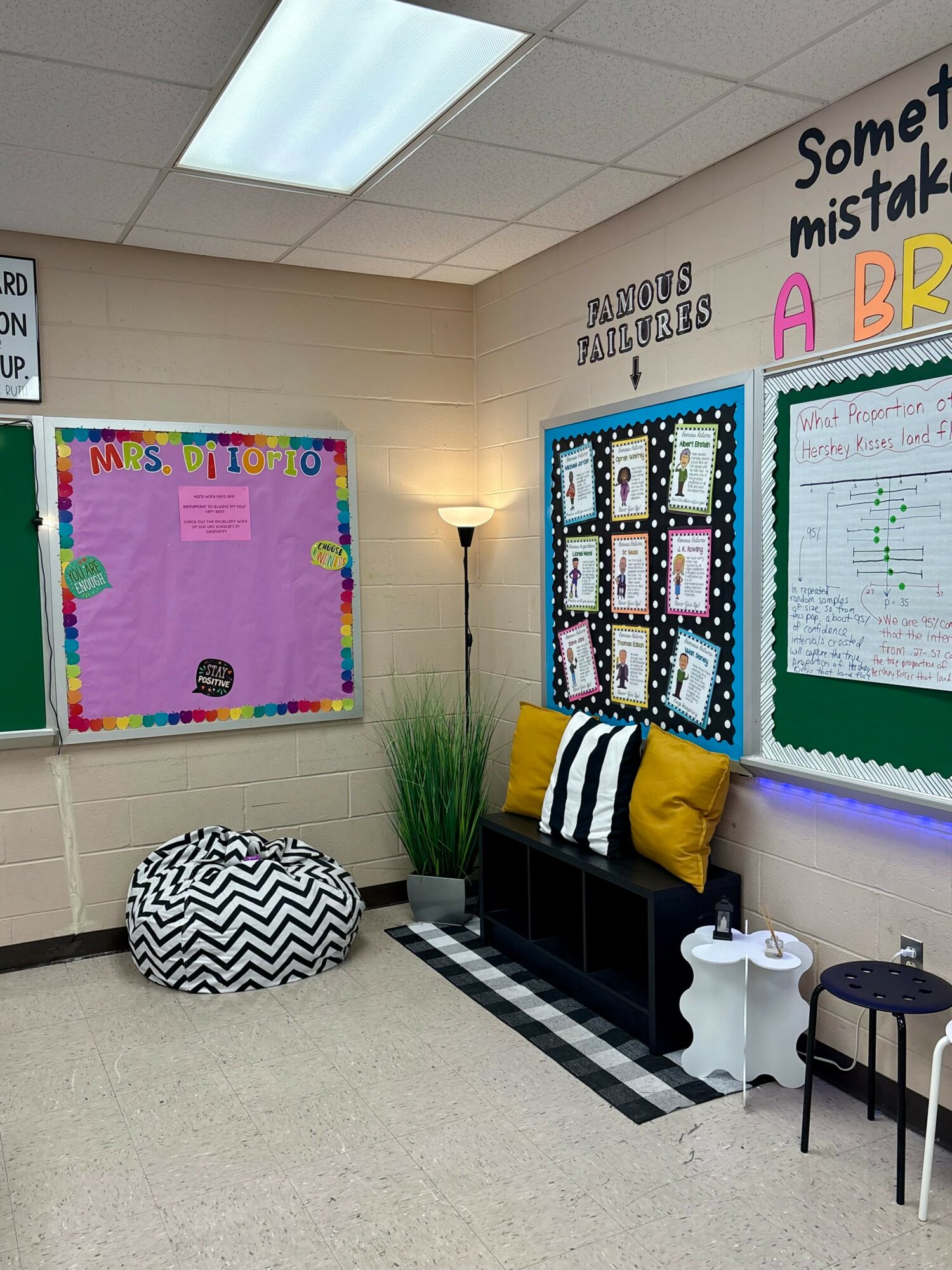 Classroom corner with bulletin boards, a black bench with yellow and black pillows, a white lamp, and a black and white patterned pouf.