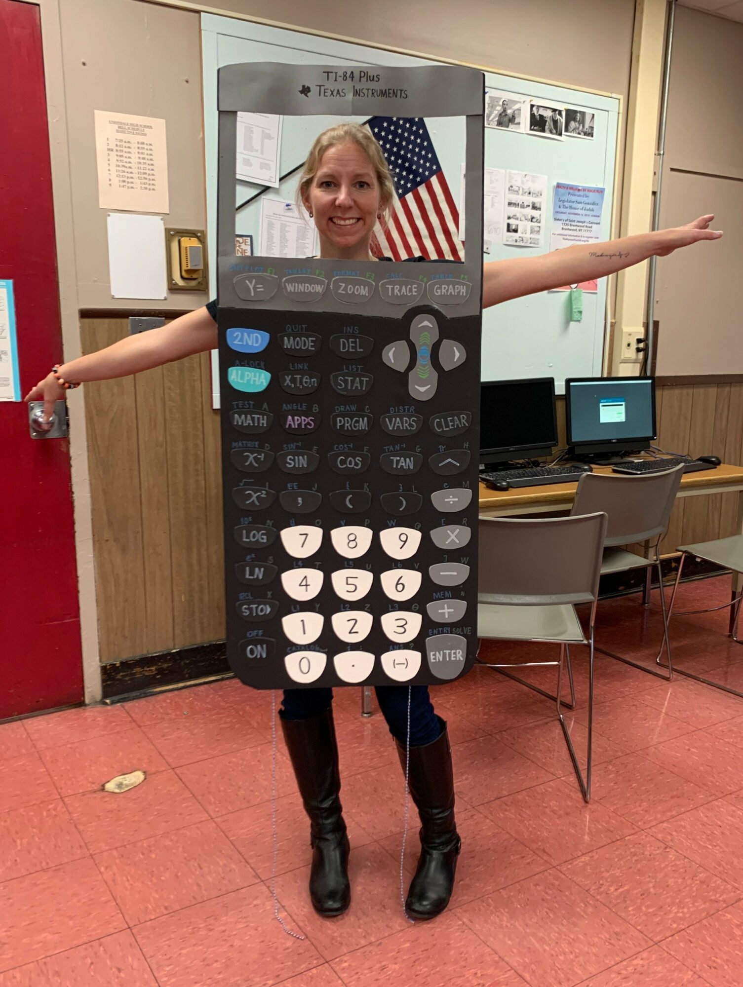 Person wearing a large calculator costume with arms outstretched, standing in a classroom or office setting.