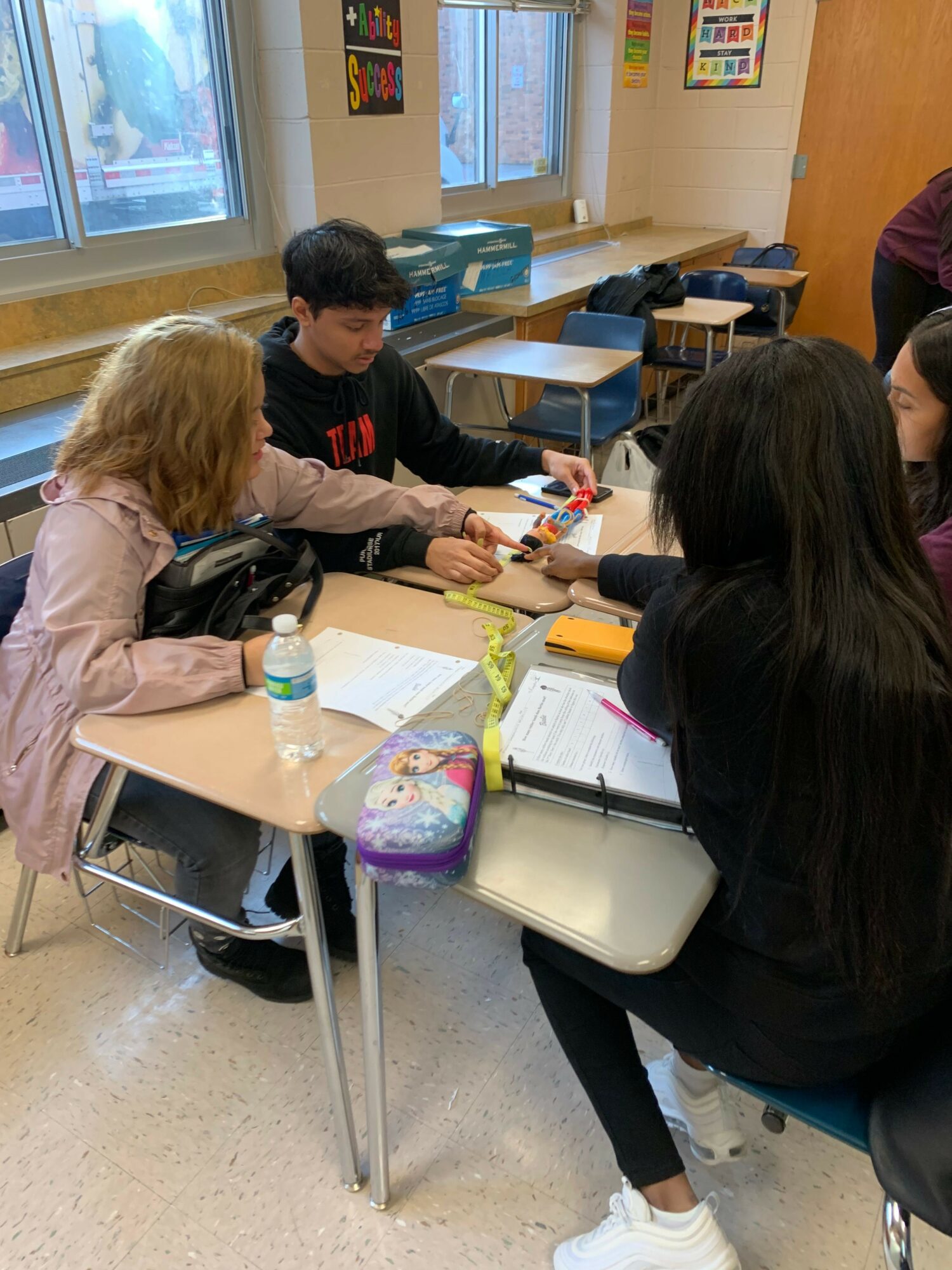 Students seated at desks in classroom, working on assignments, with books and supplies on desks.