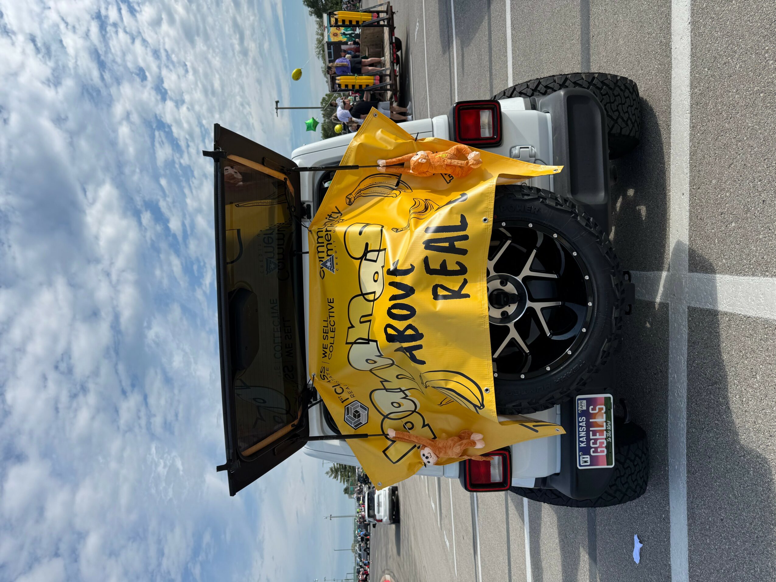 Pickup truck with a yellow banner reading 'Above the Real' attached to the back, parked in a parking lot under a cloudy sky.