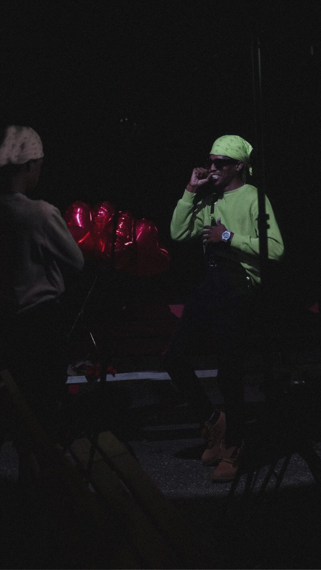 Person in bright green clothing and hat talking to another person in a dark setting, with red lights in the background.
