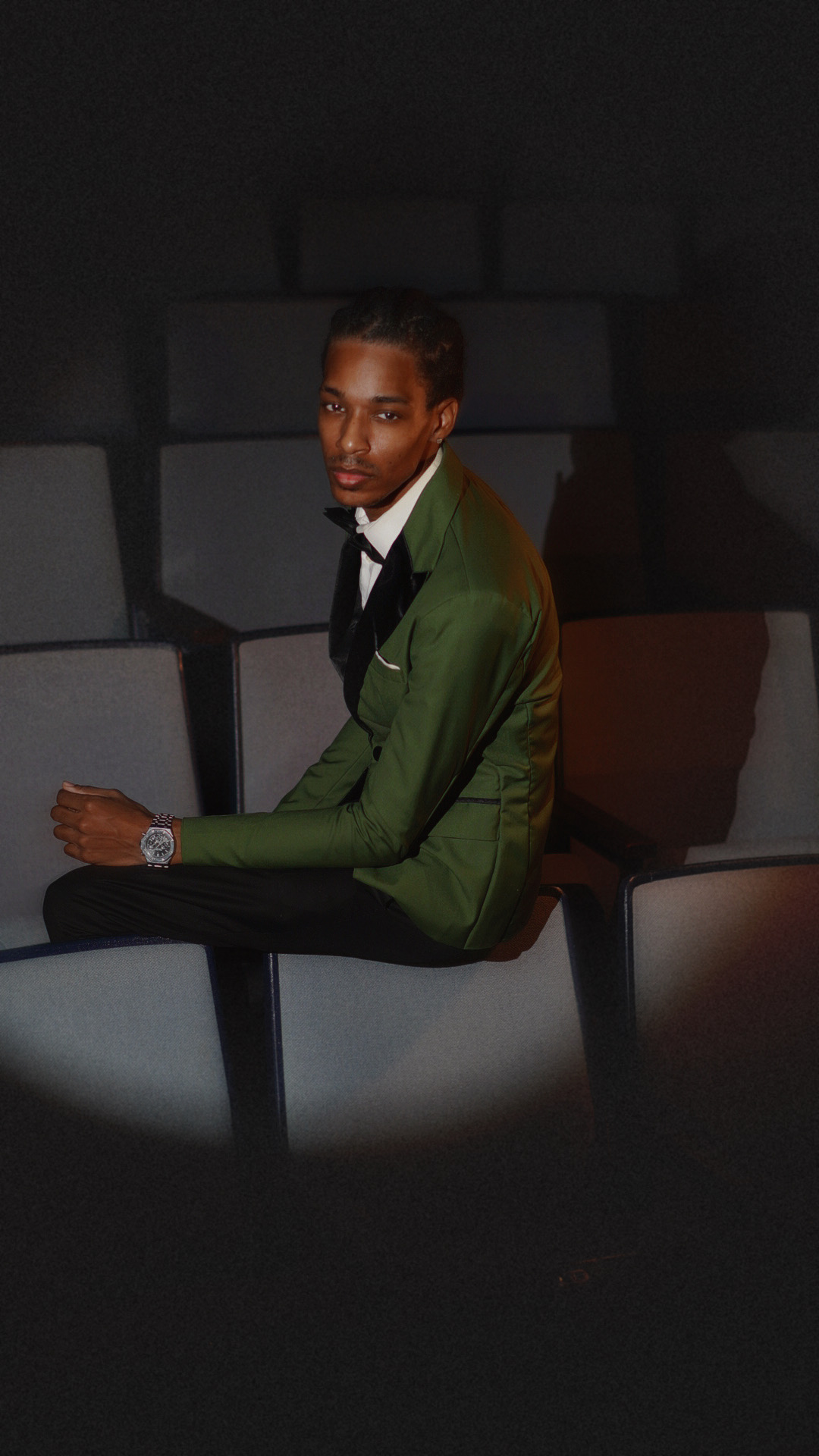 Young man in a green suit sitting in a dark room with geometric wall patterns, looking at the camera.