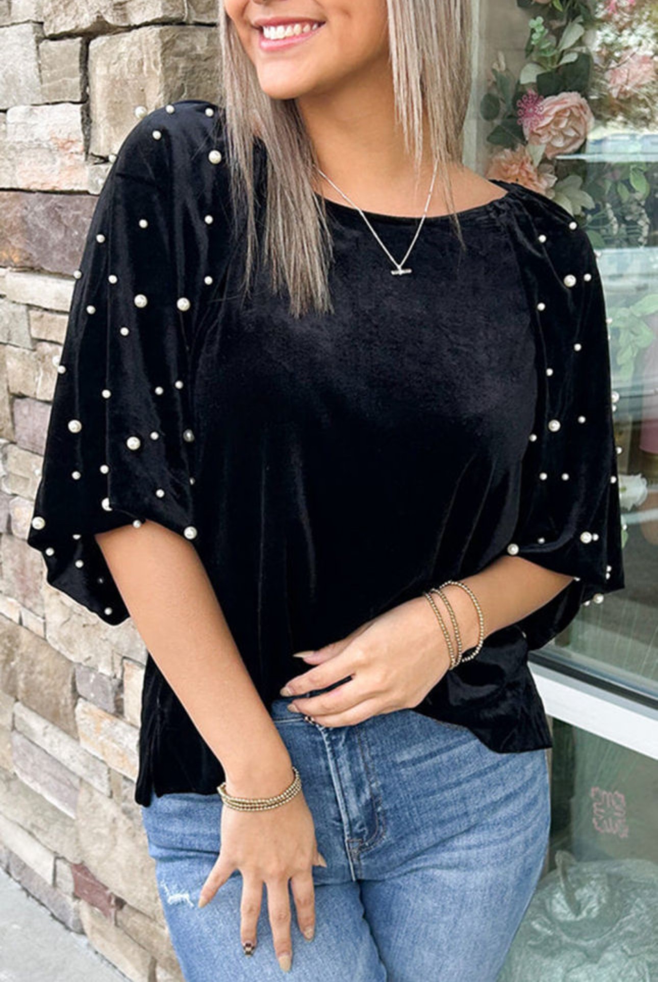 Woman wearing a black top with pearl embellishments on sleeves, smiling, standing outdoors near a brick wall and flowers.