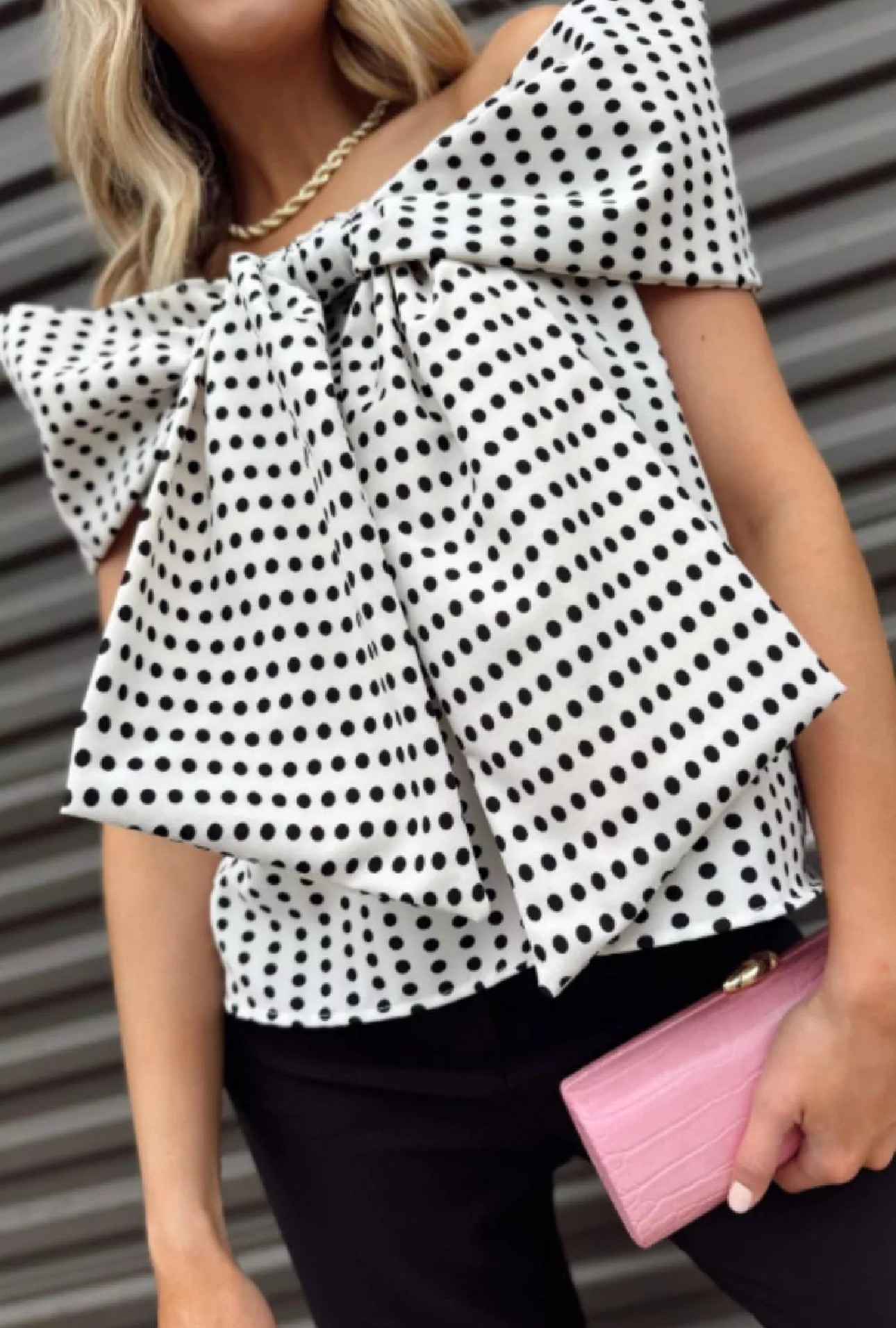 Person wearing a polka dot top with a large bow, holding a pink clutch, standing in front of a gray background.