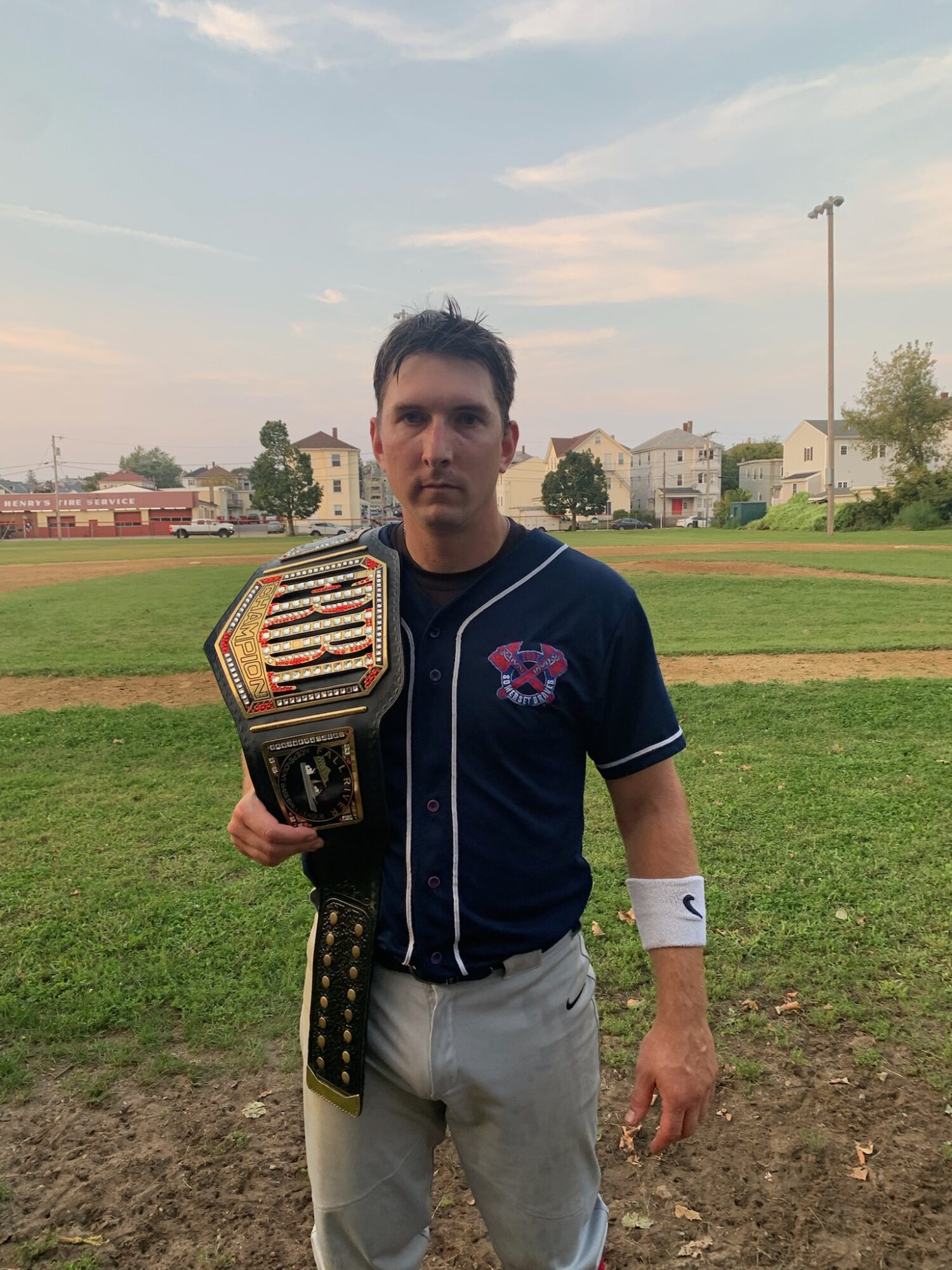 Young man in baseball uniform holding a championship belt on a grassy field with houses in the background.