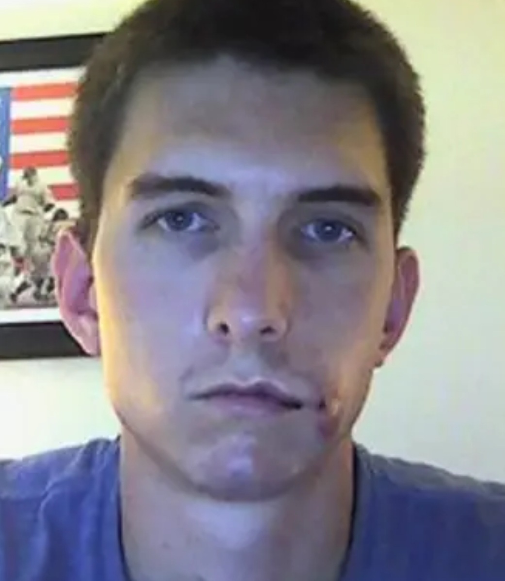 Young man with short dark hair and blue eyes, neutral expression, wearing a blue shirt, background with an American flag picture.