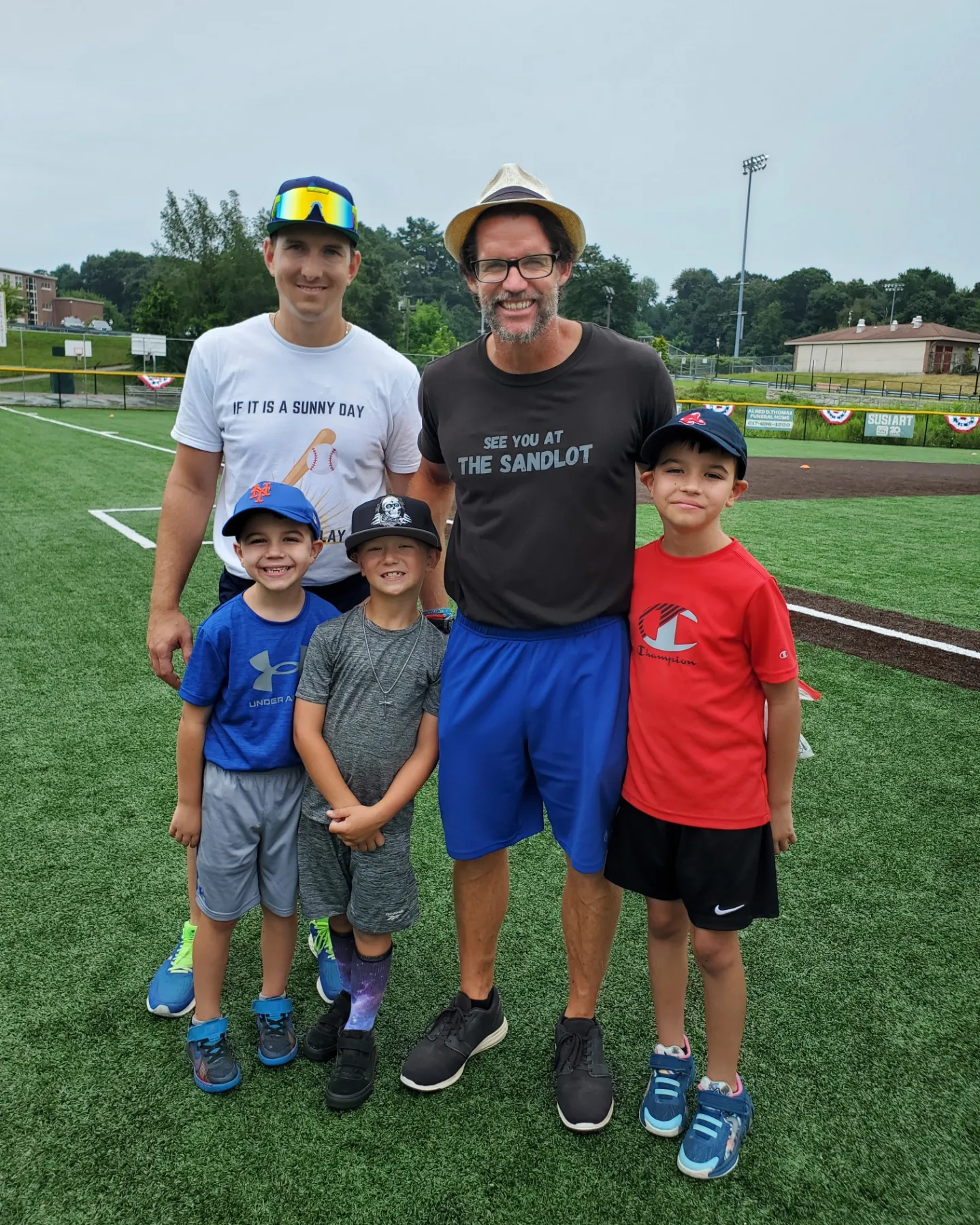 Group of five people, including two adults and three children, standing on a grassy field with a sports field in the background.
