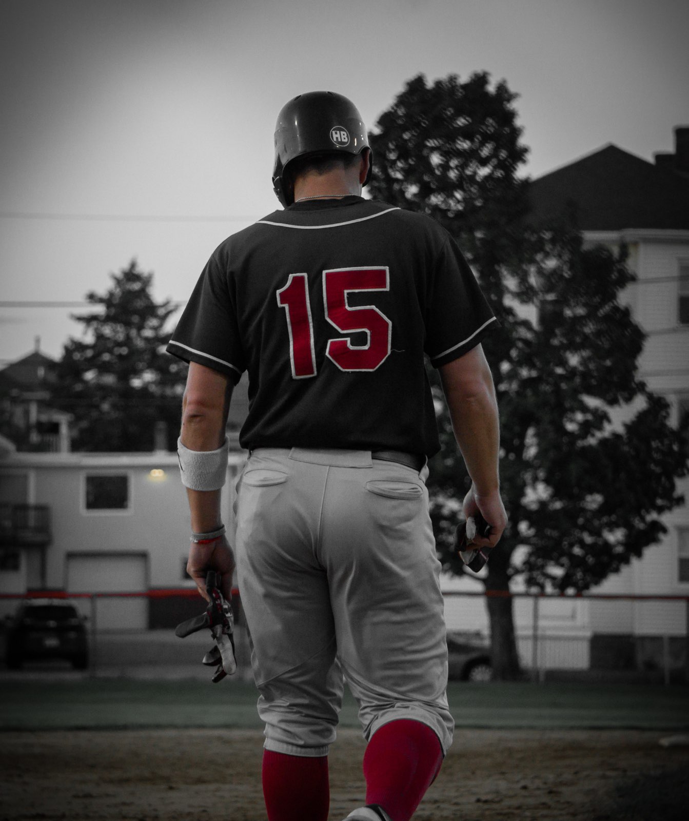 Baseball player standing on field with trees and buildings in background, wearing helmet, jersey number 15, gray pants, and red socks.