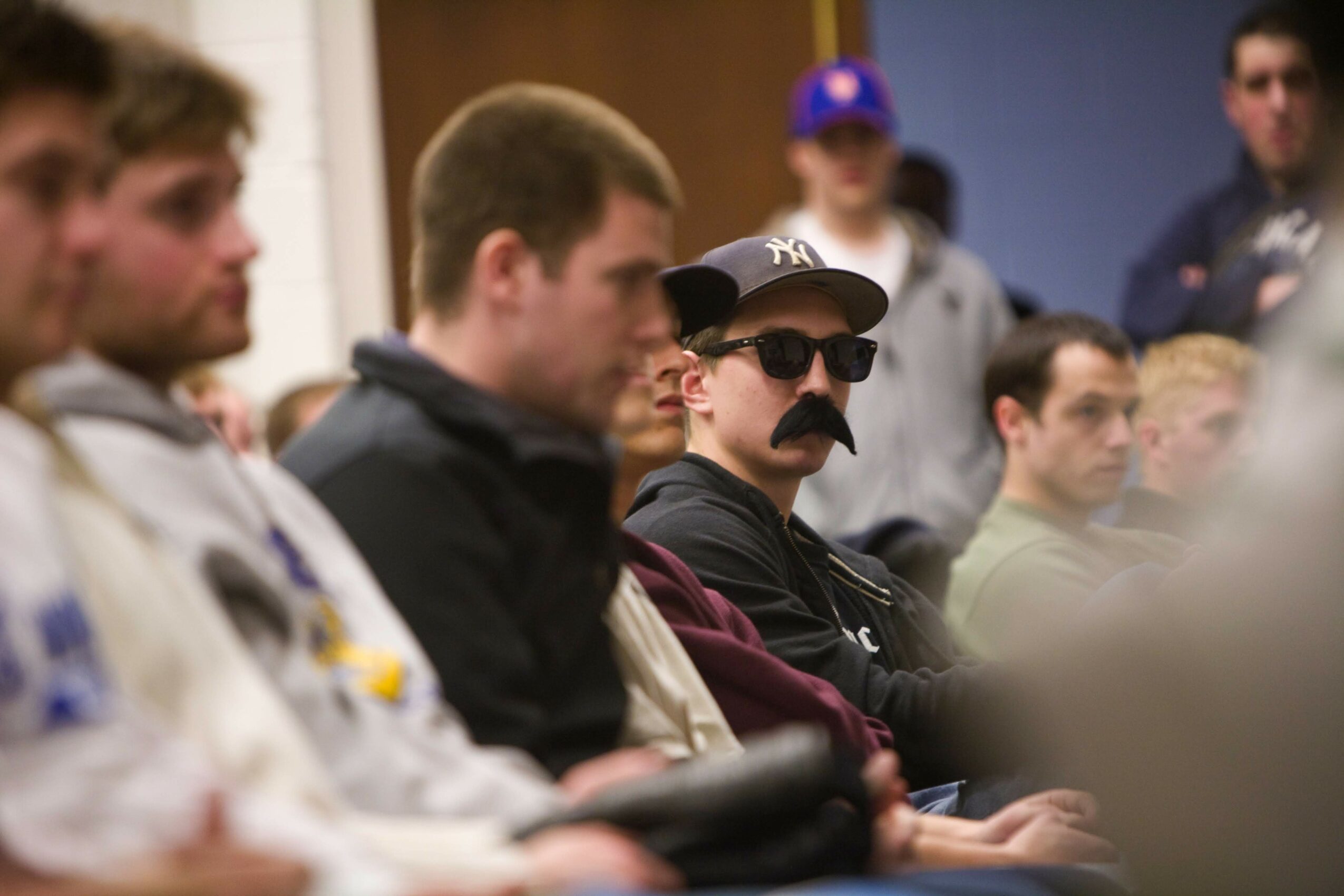 Person wearing sunglasses and a fake mustache, sitting in a room with other people, some standing and some seated.