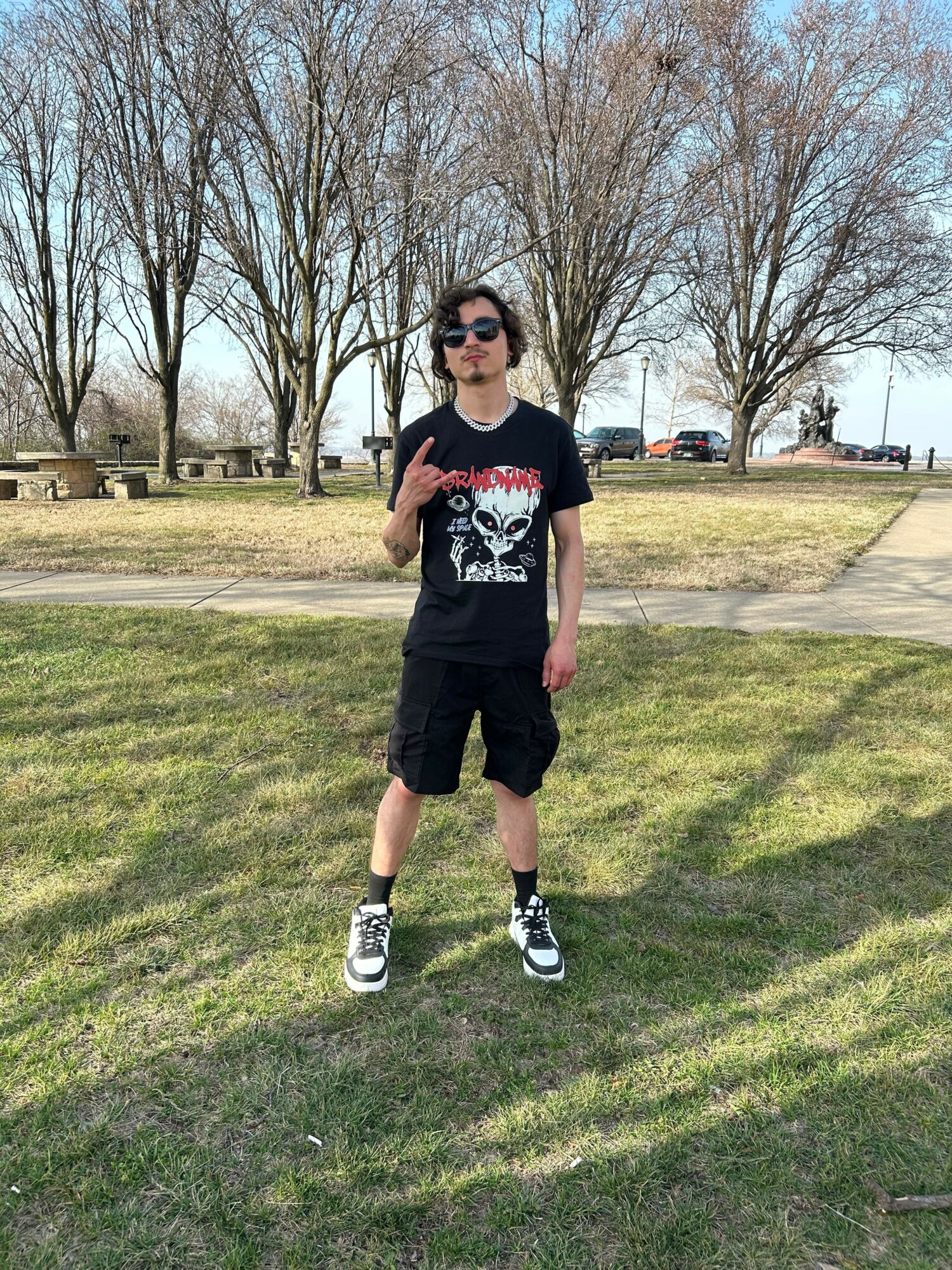 Young person standing on grass in park, wearing sunglasses, black t-shirt with skull design, black shorts, and sneakers.
